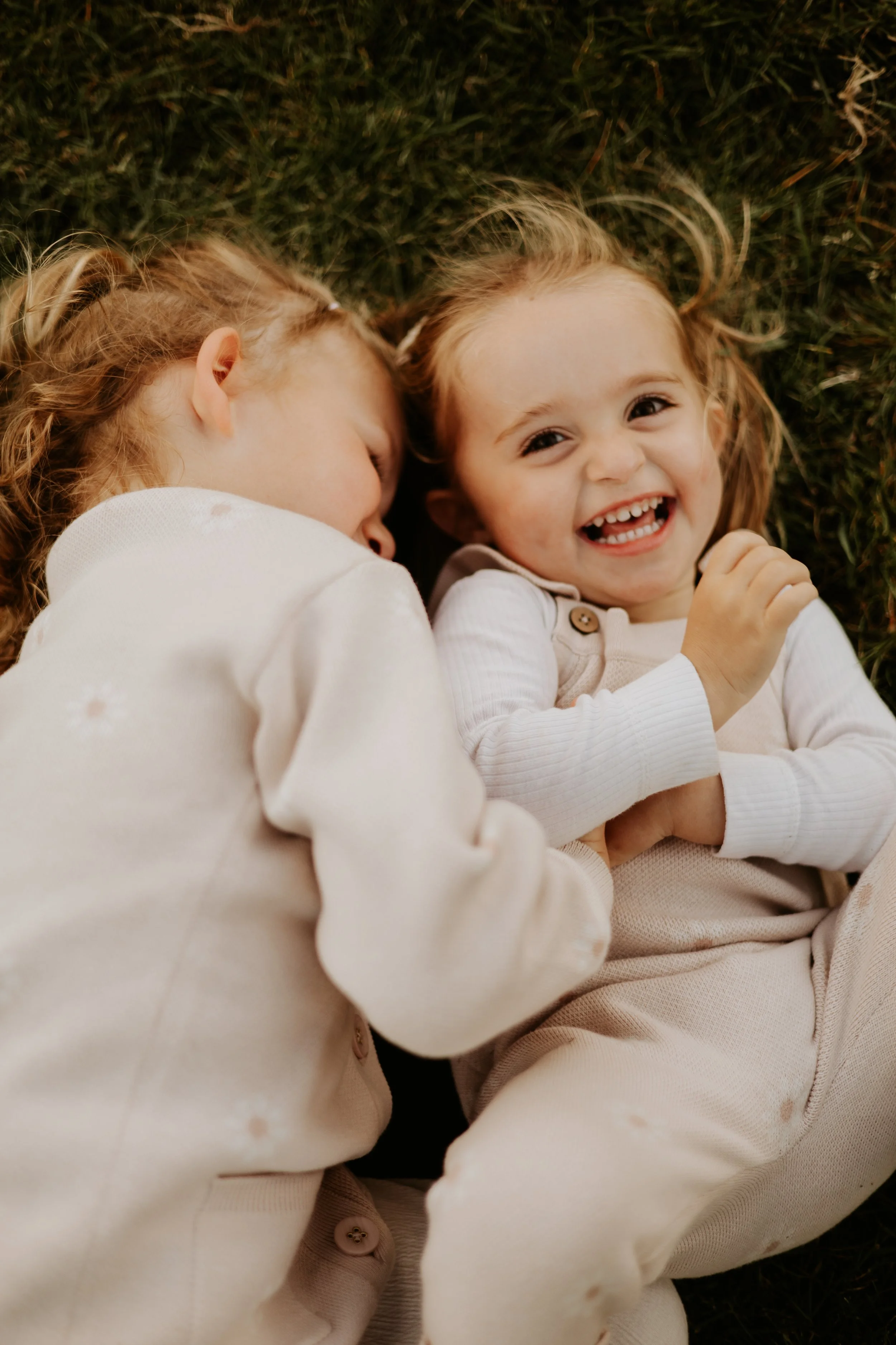 Two young girls with light skin and red hair lying on the grass, one is asleep while the other is smiling and laughing.