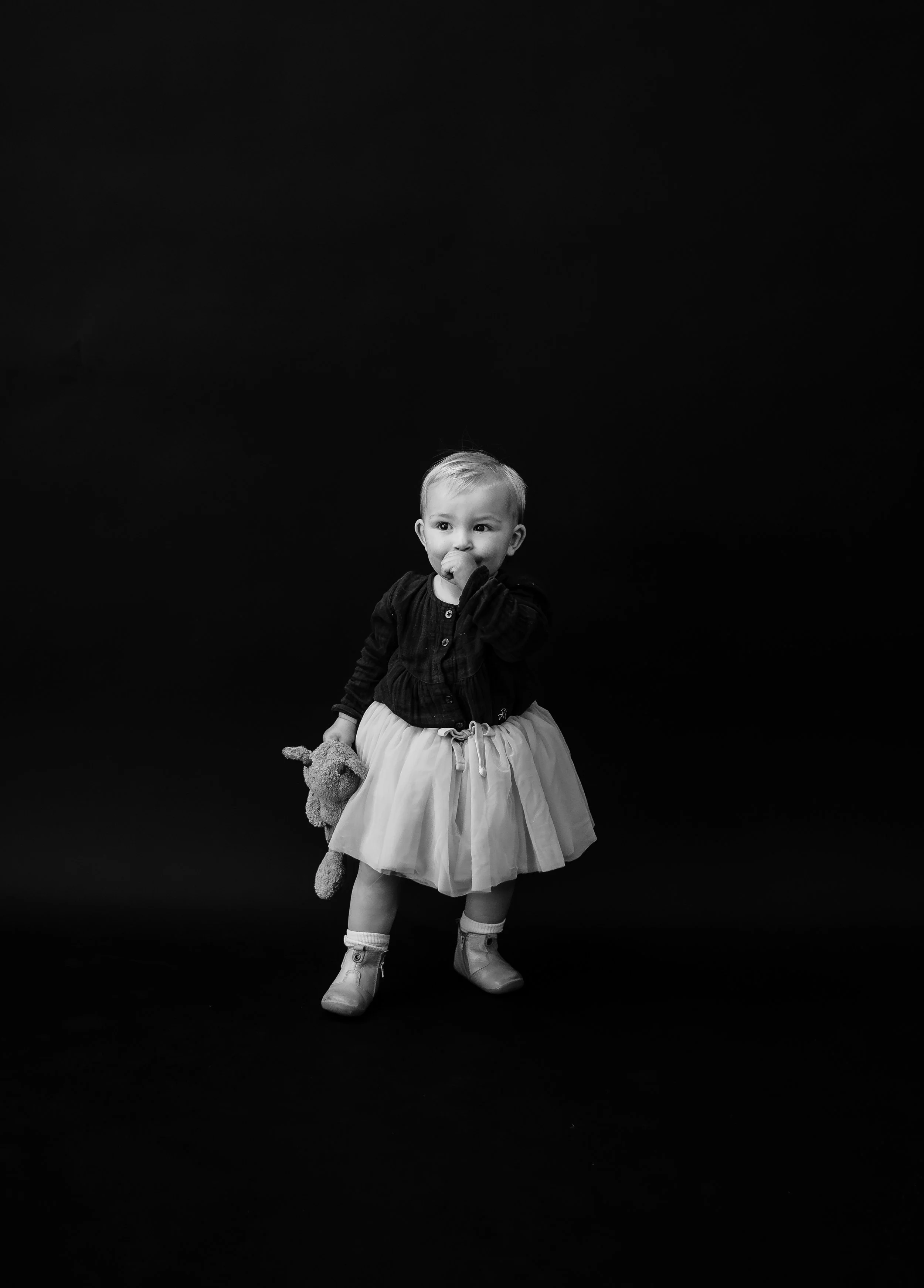 A young girl standing against a black background, holding a stuffed animal in her left hand, wearing a light-colored skirt, a dark top, and boots, with one hand near her mouth.