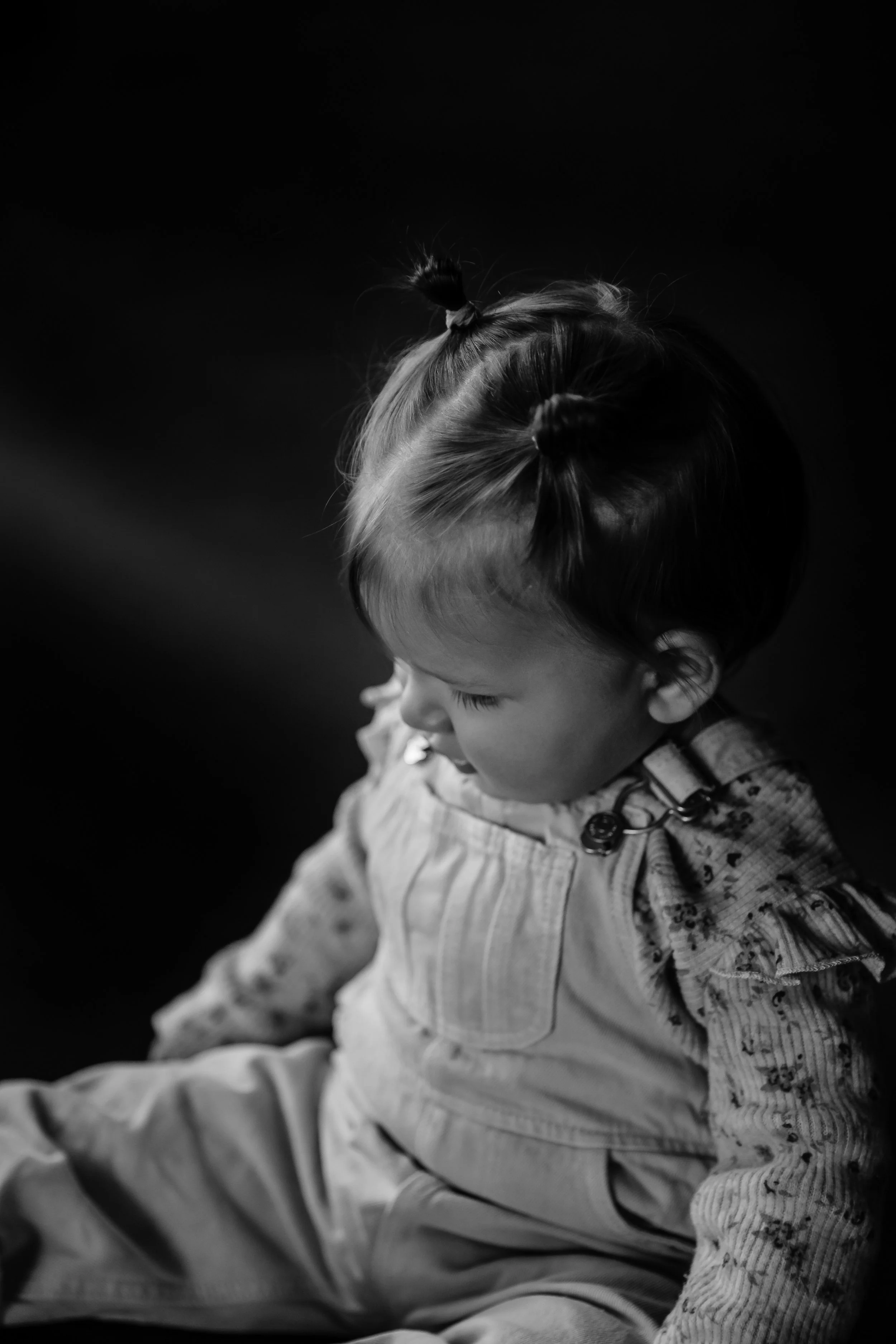 A young girl with two small buns in her hair, looking downward, sitting against a dark background.