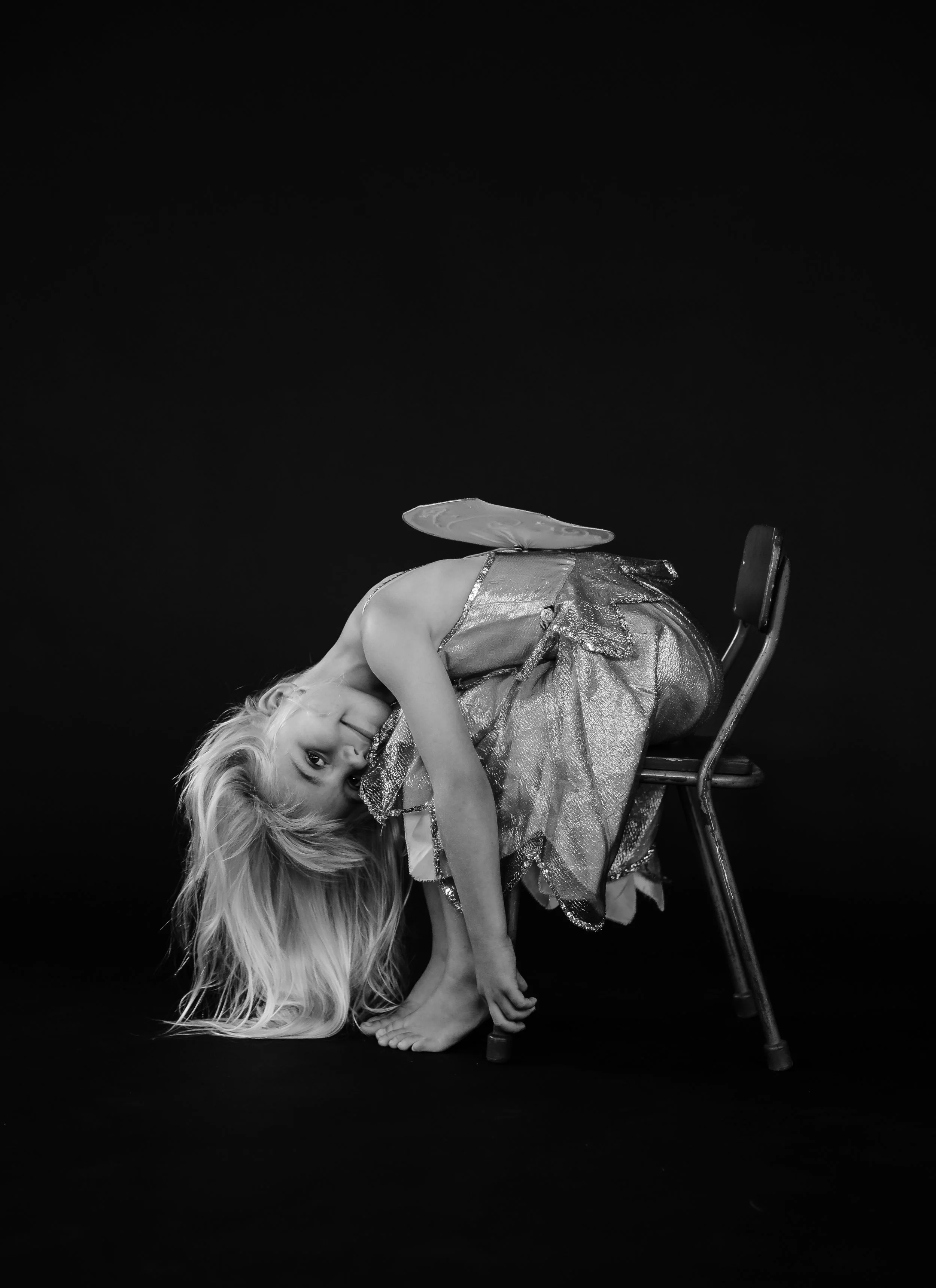 A girl with long blonde hair bending backwards on a chair, wearing a metallic dress and fairy wings, against a black background.