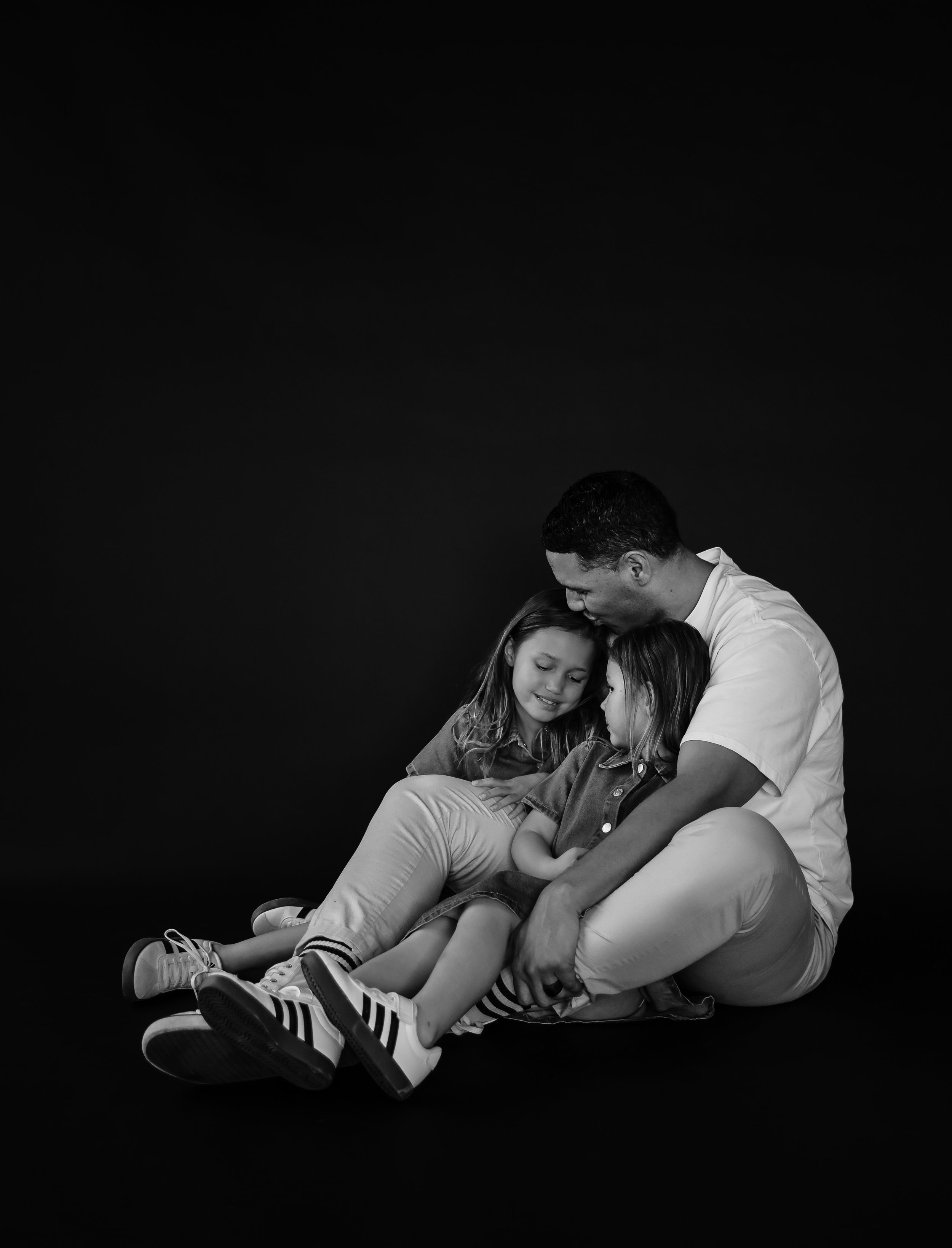 Black and white photo of a man with two young girls sitting on the floor, hugging and smiling, against a dark background.