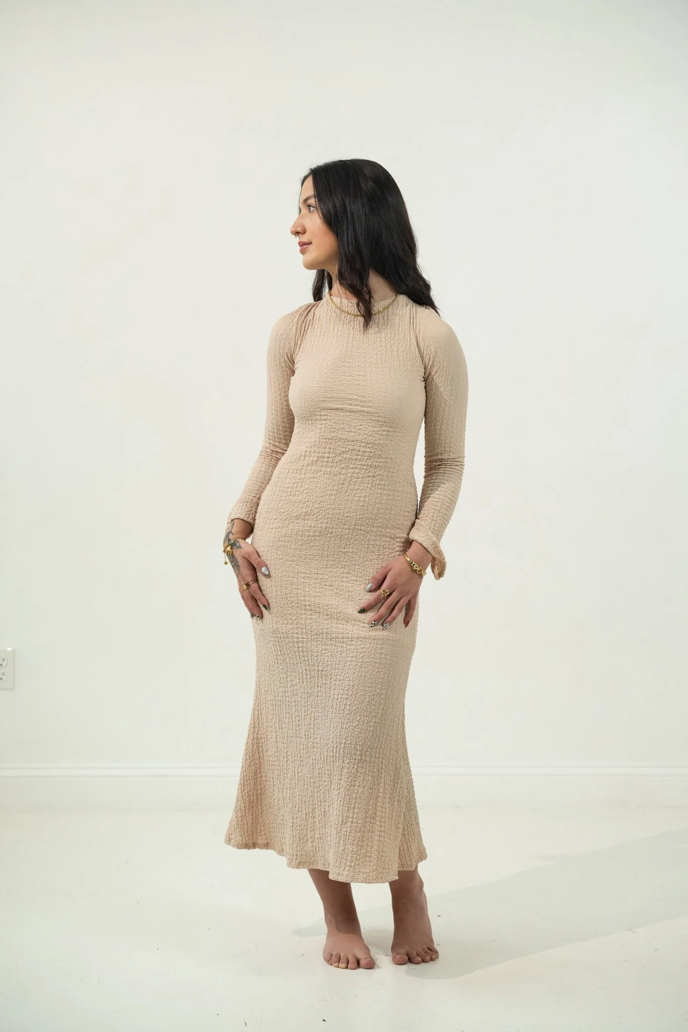 A woman with long dark hair wearing a beige long-sleeve textured dress standing barefoot against a plain white wall.