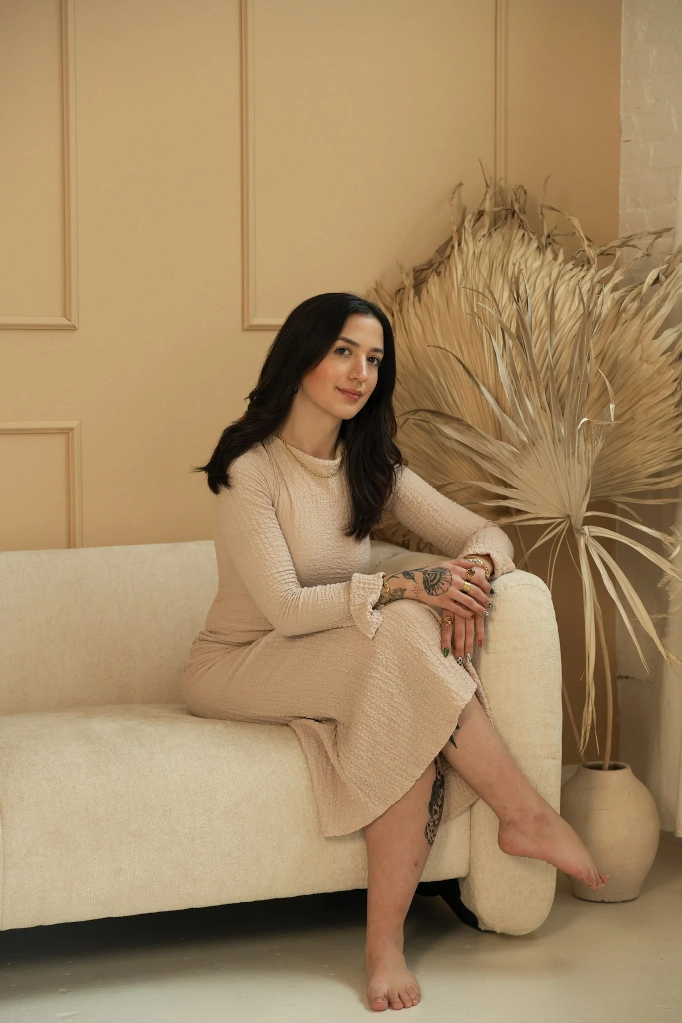 A woman with dark hair sitting on a cream-colored couch in a beige dress, with tattoos on her arm and leg, in a room with beige walls and a large dried palm leaf in a vase.
