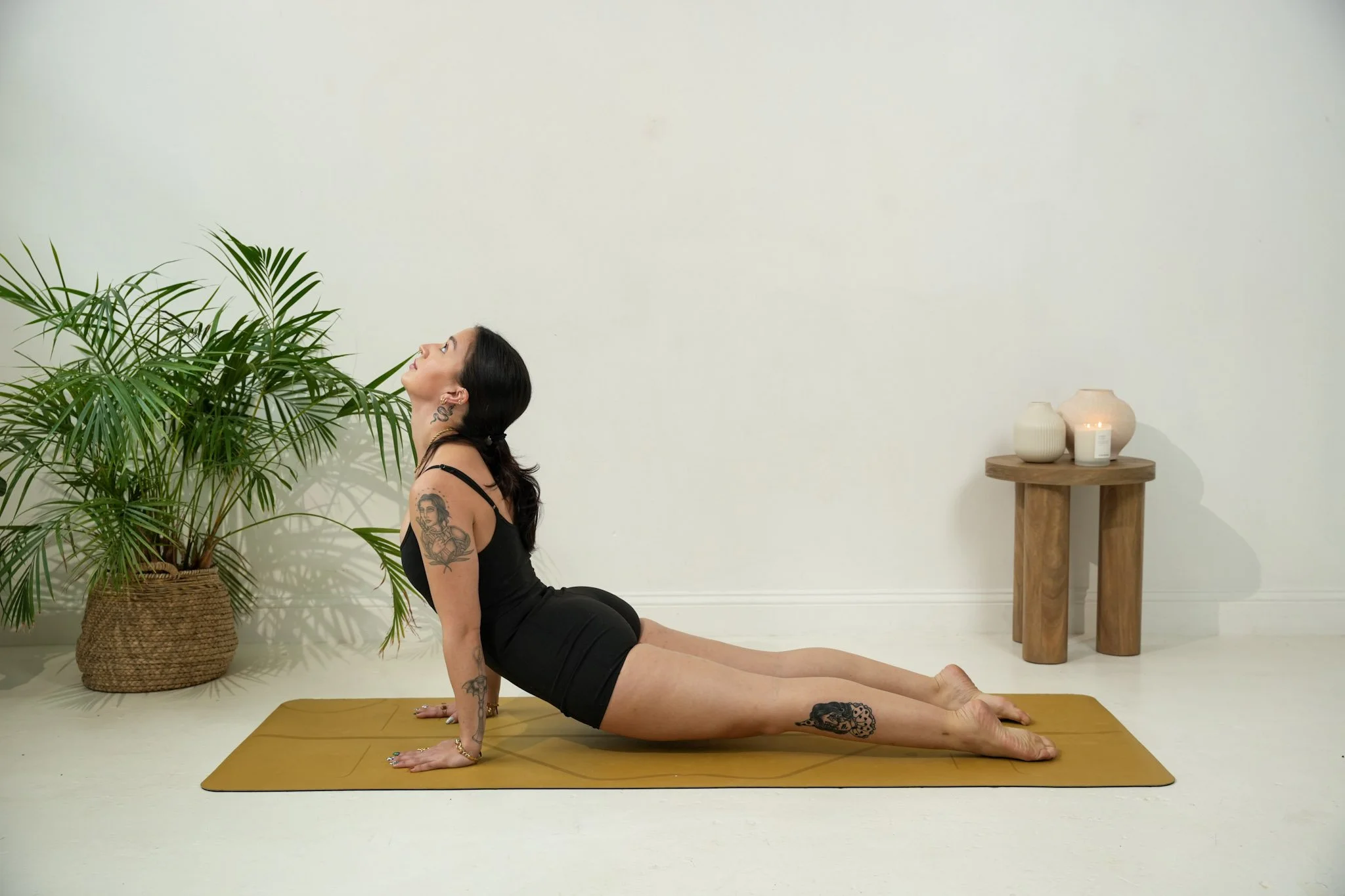 Woman in black workout outfit performing a yoga pose on a yoga mat in a minimalistic room with potted plants and candles.