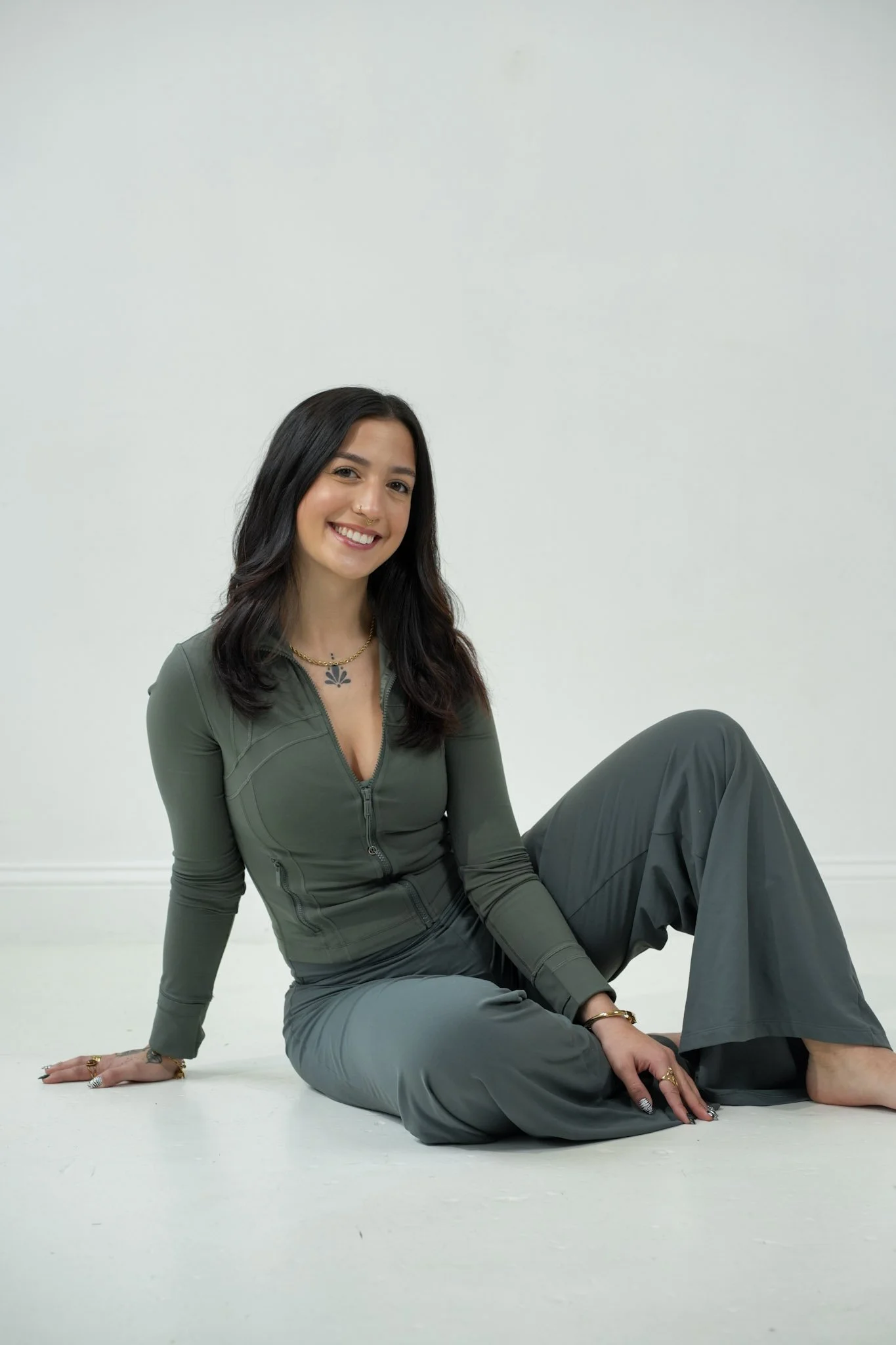 A woman with dark hair, smiling, sitting on a white floor against a plain white wall, dressed in a green zip-up top and wide-leg gray pants.