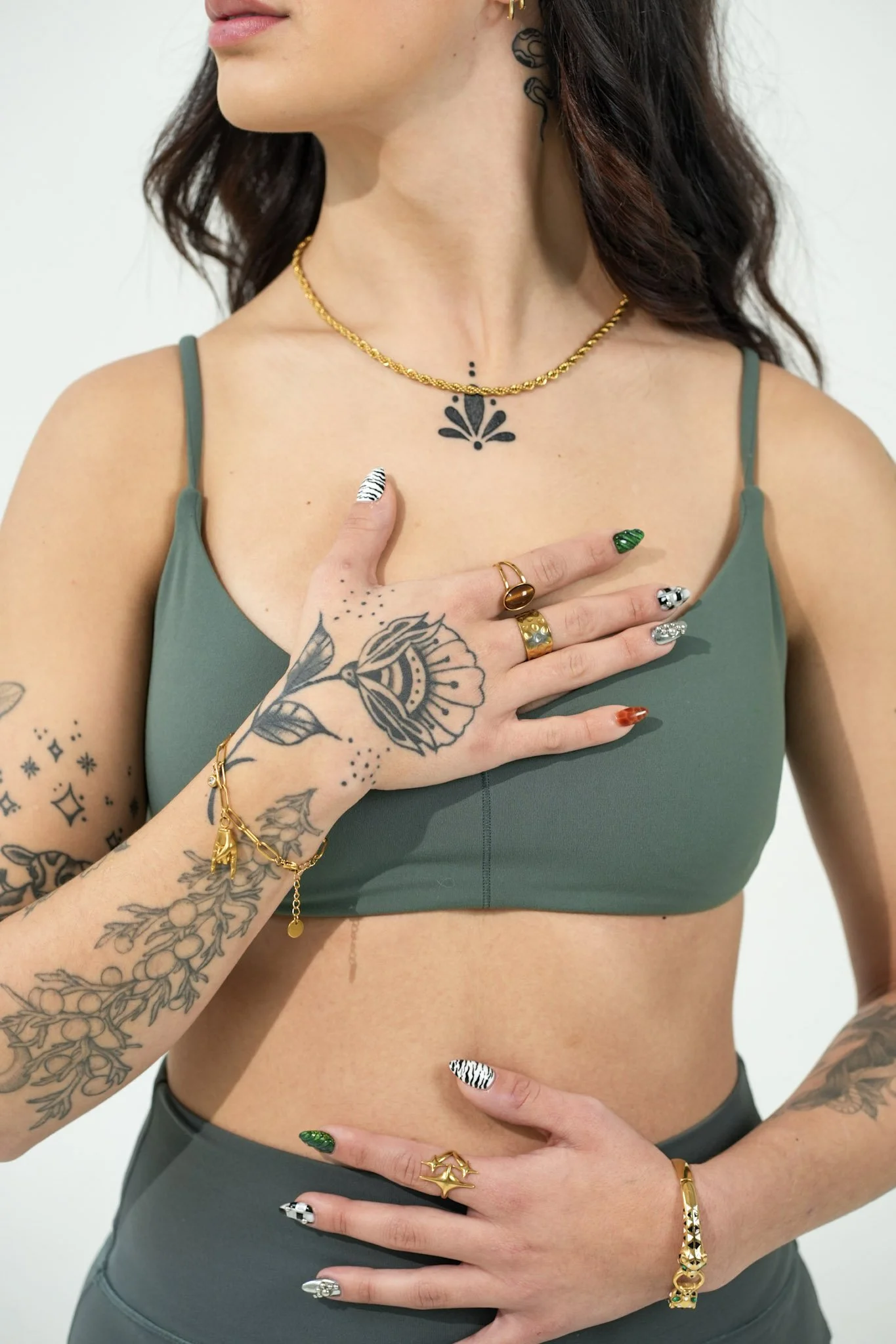 Close-up of a woman's torso, showing her wearing a green sports bra, multiple gold rings, bracelets, and necklaces, with tattoos on her right arm, left hand, and chest, and her hand placed over her stomach.