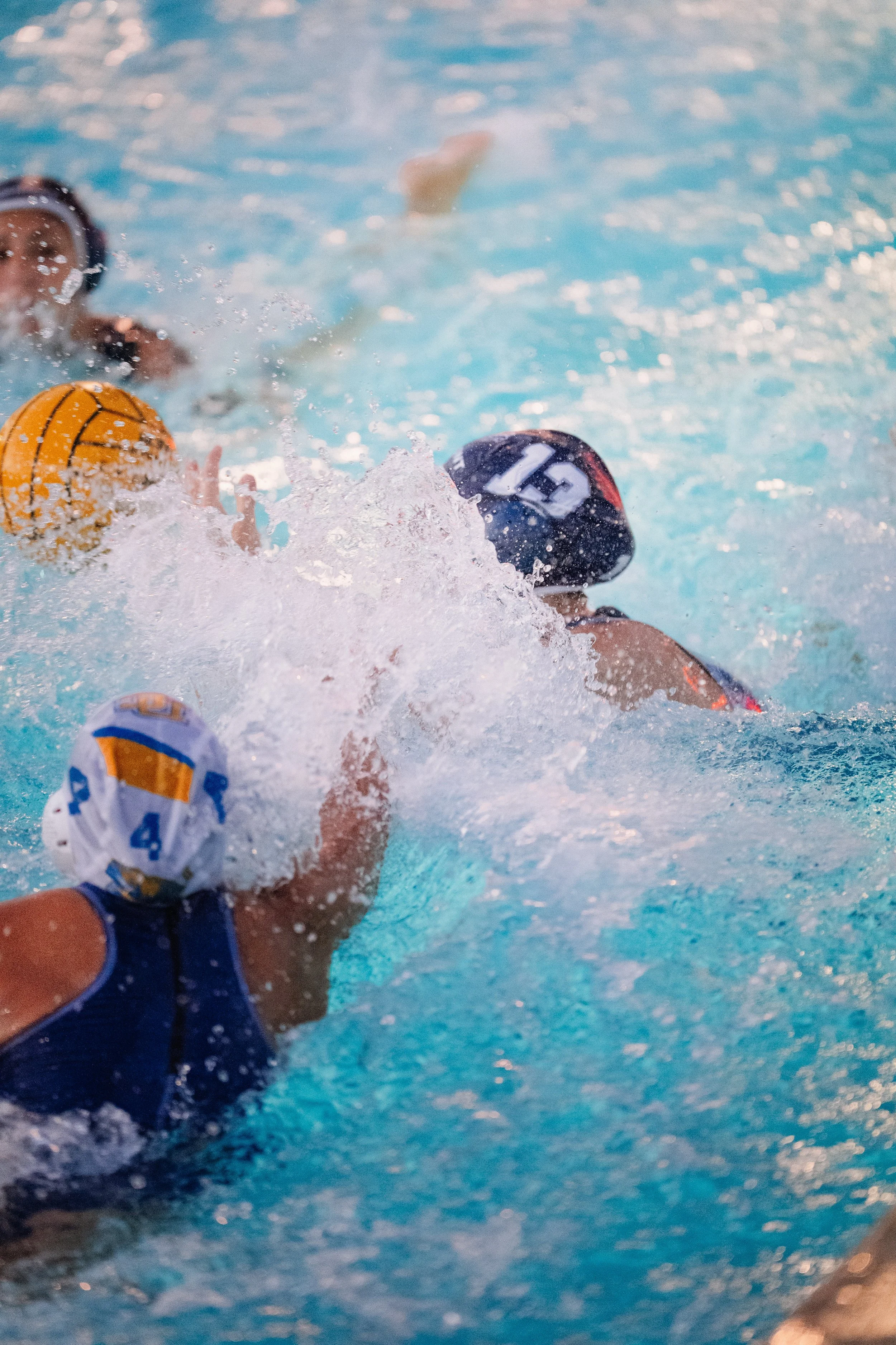Water polo players during a match, with one player holding a yellow ball and another wearing a cap with the number 12, in a swimming pool with splashing water.
