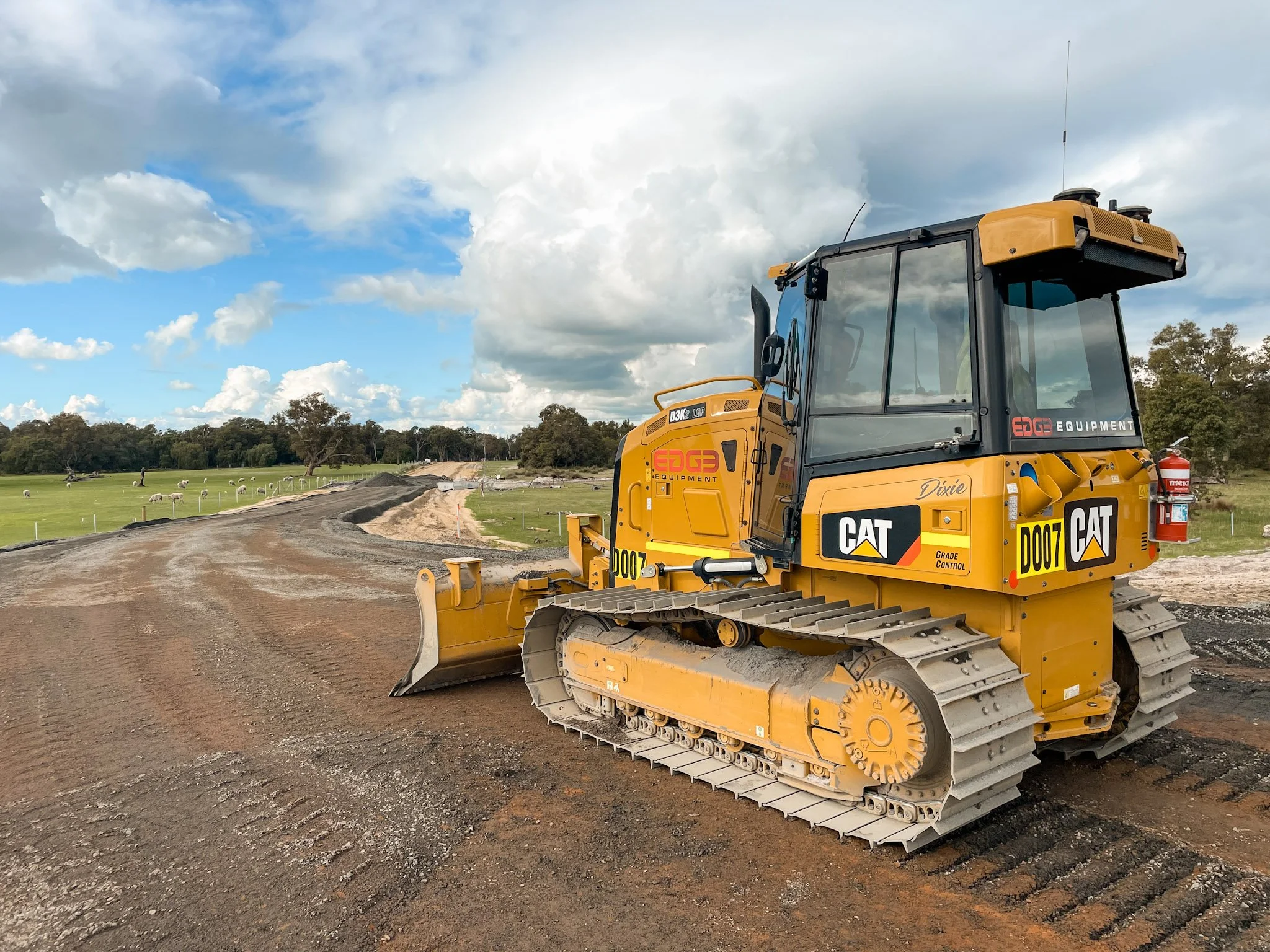 A yellow CAT bulldozer working on a dirt road construction site under a partly cloudy sky, with green fields and trees in the background.