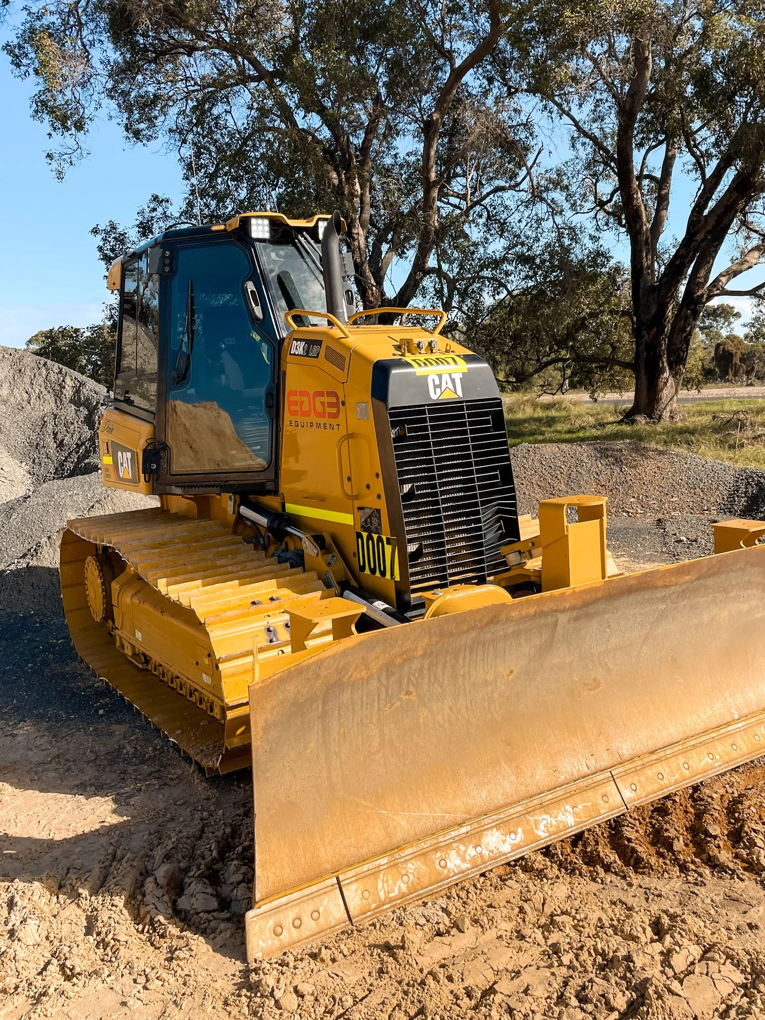 A yellow Caterpillar bulldozer with a large blade at the front, on a dirt construction site with trees in the background.