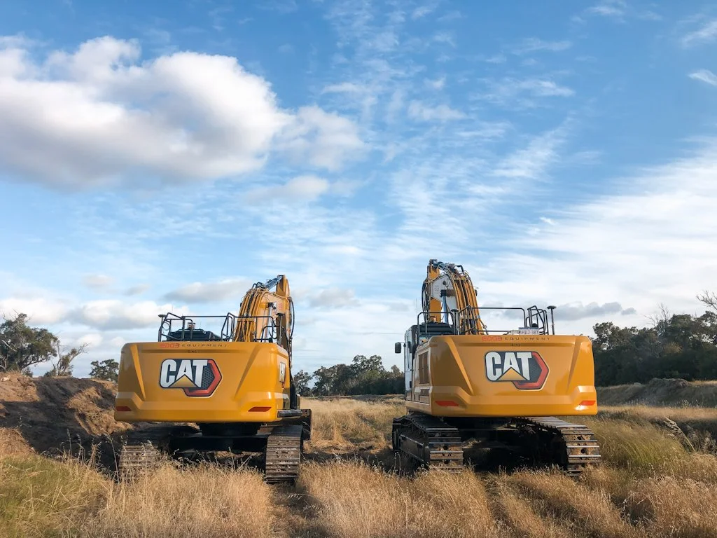 Two yellow Caterpillar excavators on a grassy field with trees in the background under a partly cloudy sky.