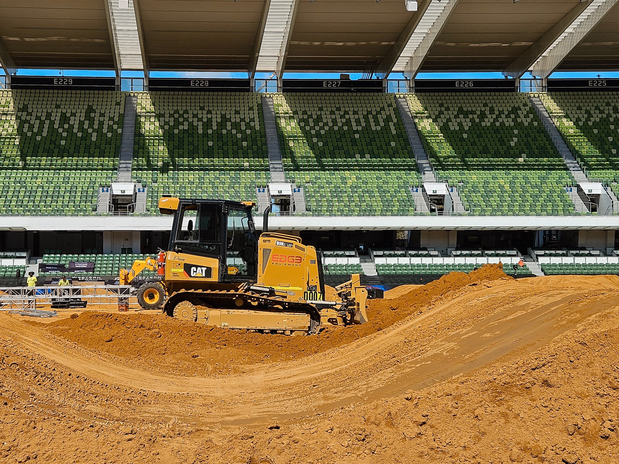 A yellow Caterpillar bulldozer grading dirt on a sports stadium field with green seating in the background.