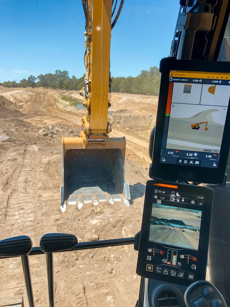 Inside the cab of a construction excavator, view of the digging bucket and a dirt construction site ahead with trees in the distance.