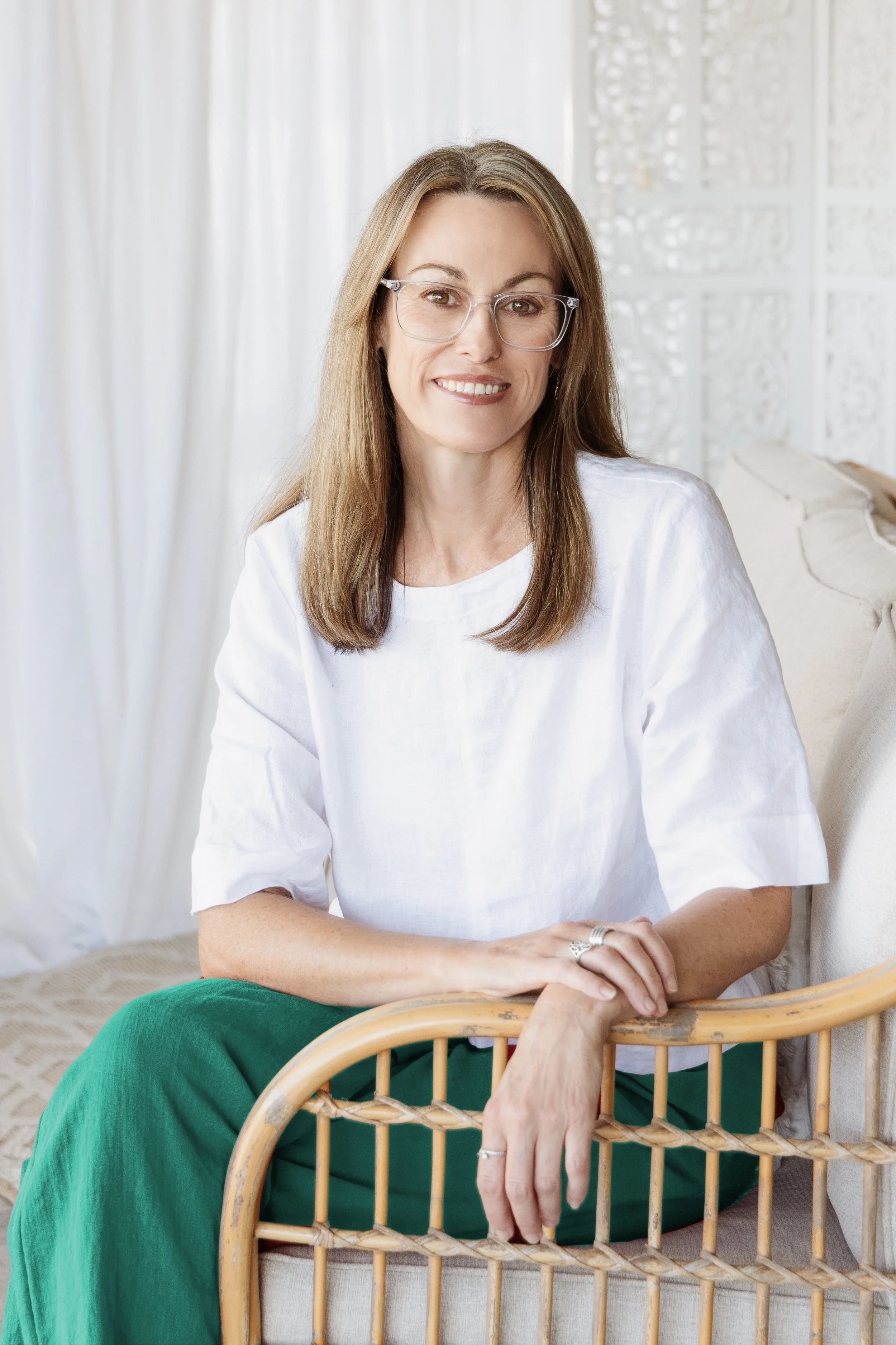 Lisa Proctor, long brown hair, glasses, white top and green pants, seated on a light‑colored chair in a bright room – representing Blue Sky Planning & Environment’s town‑planning and environmental‑consulting expertise.