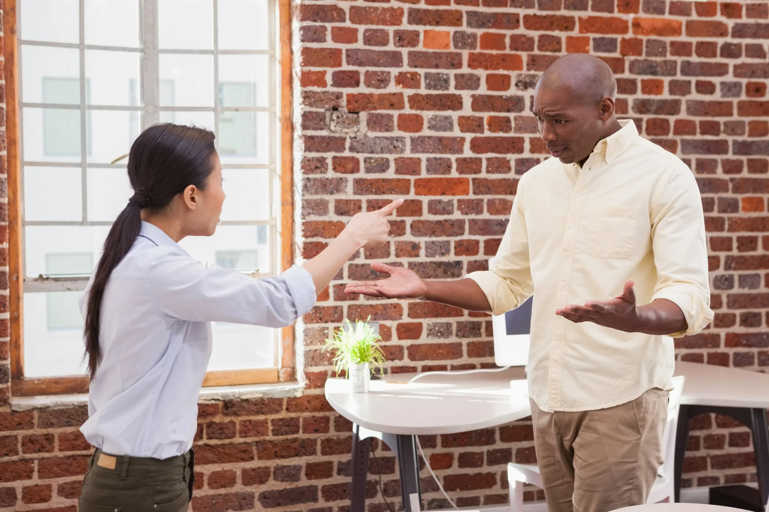 A woman with dark hair in a ponytail pointing at a man with a gesture of confusion or disagreement in front of a brick wall and large window in an office.