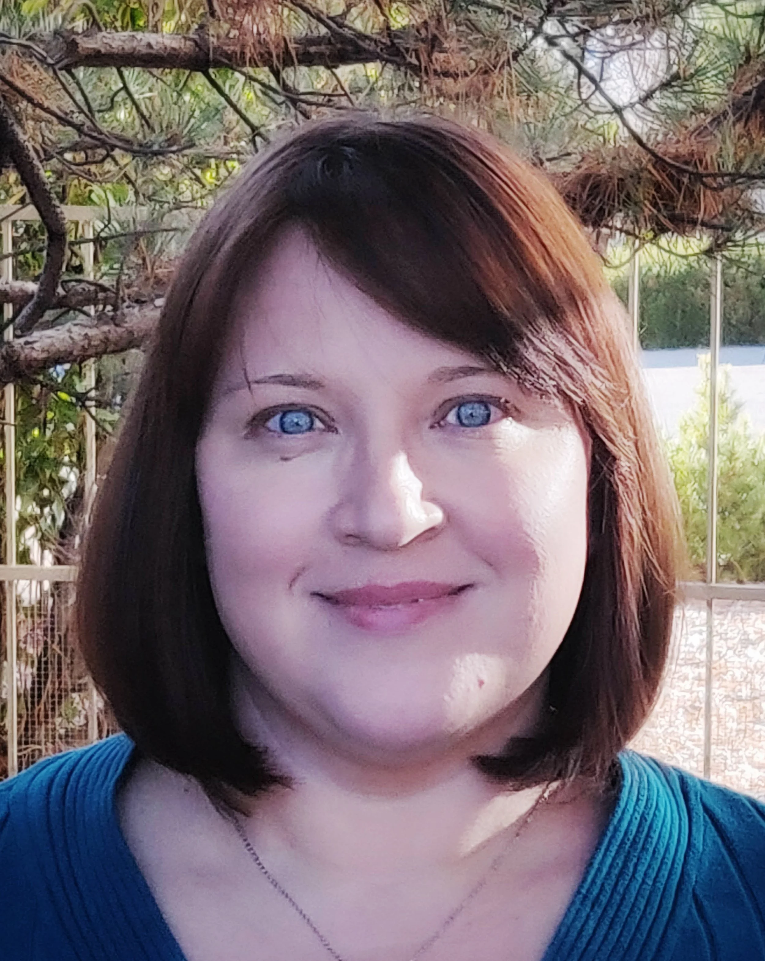 Close-up of a woman with shoulder-length brown hair and blue eyes, smiling, outdoors with trees and a garden fence in the background.