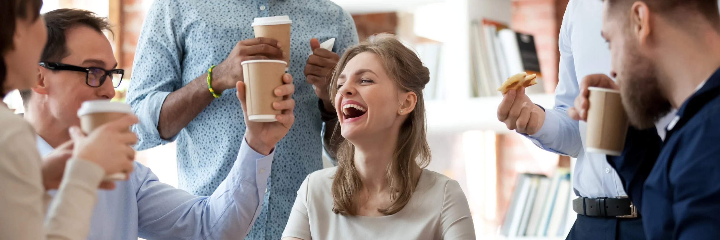 Group of people laughing and talking while holding coffee cups in a bright, modern space.