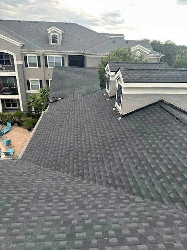 Worker repairing a brown shingled roof using tools and safety equipment, with trees and blue sky in the background.