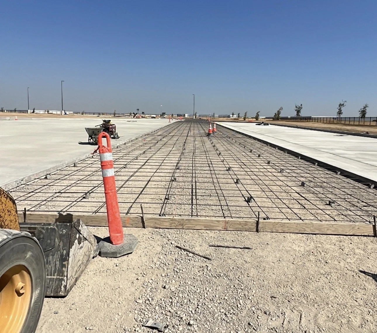 Construction site with rebar and poured concrete, perhaps preparing a road or pavement, under a clear blue sky with trees in the distance.
