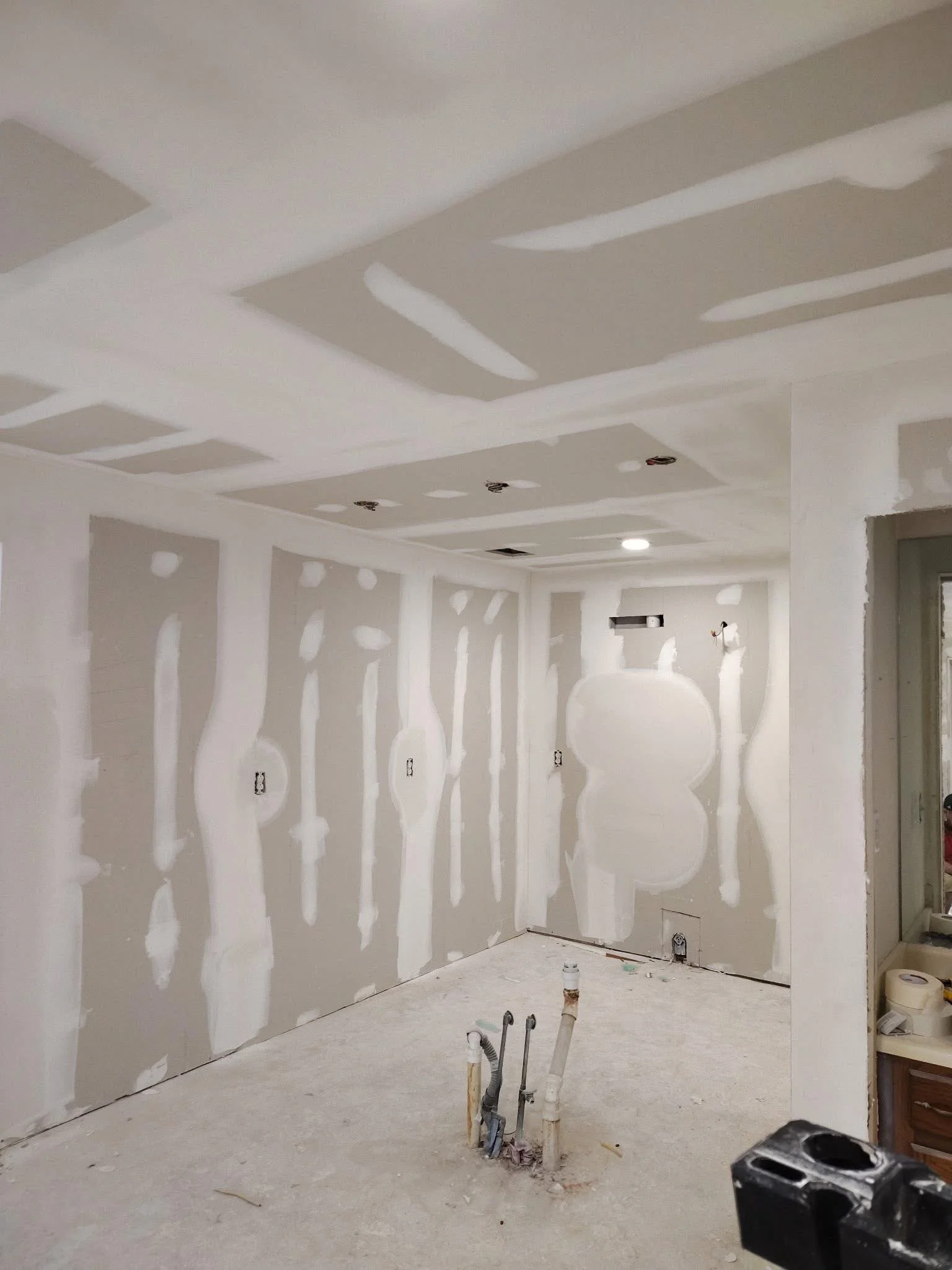 Interior of a room under construction with drywall installed and taped, no flooring, with exposed pipes and electrical outlets.