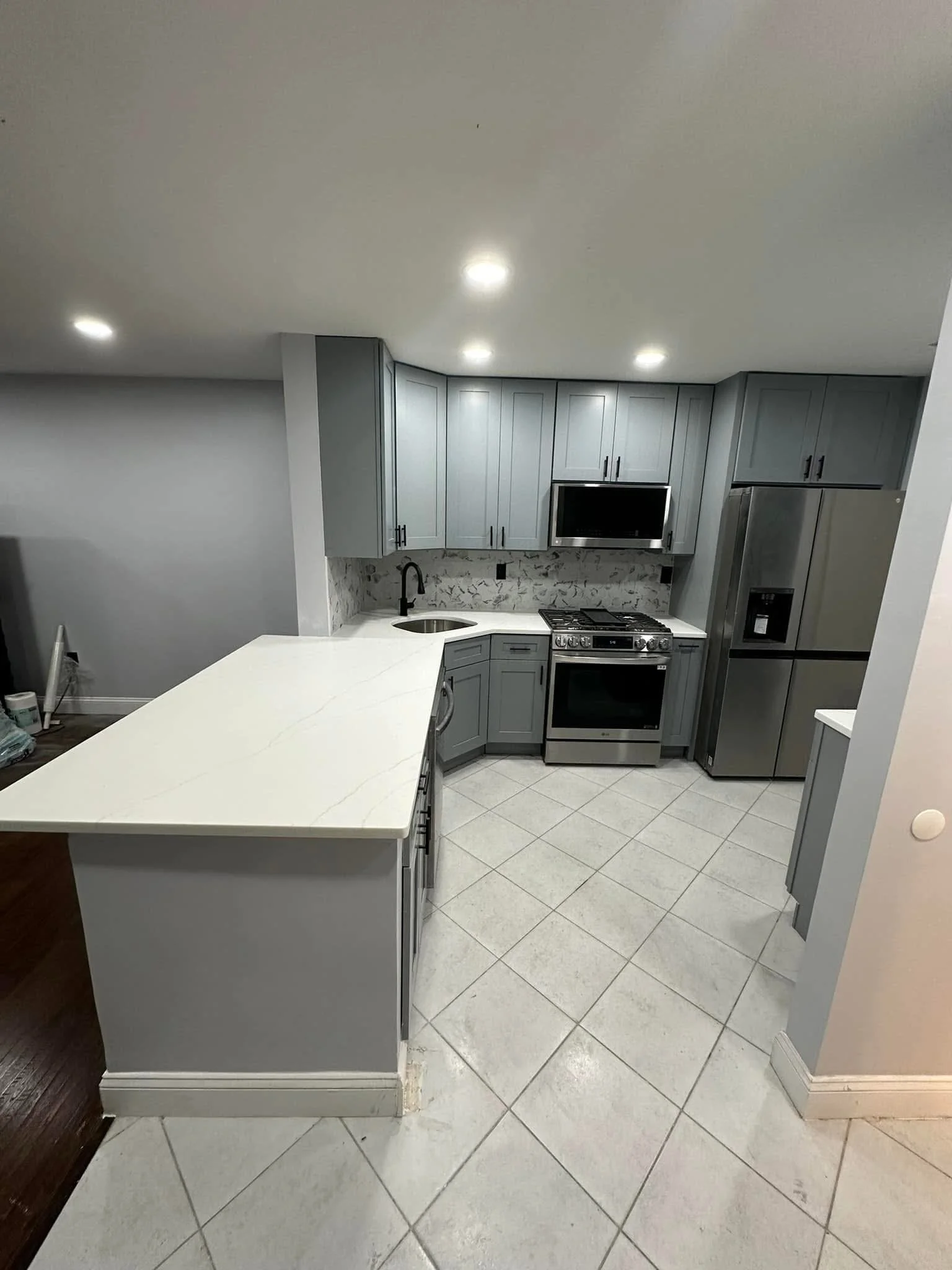 Modern kitchen with gray cabinets, white marble countertops, stainless steel refrigerator, oven, microwave, and a small sink, with tiled floor and recessed ceiling lights.