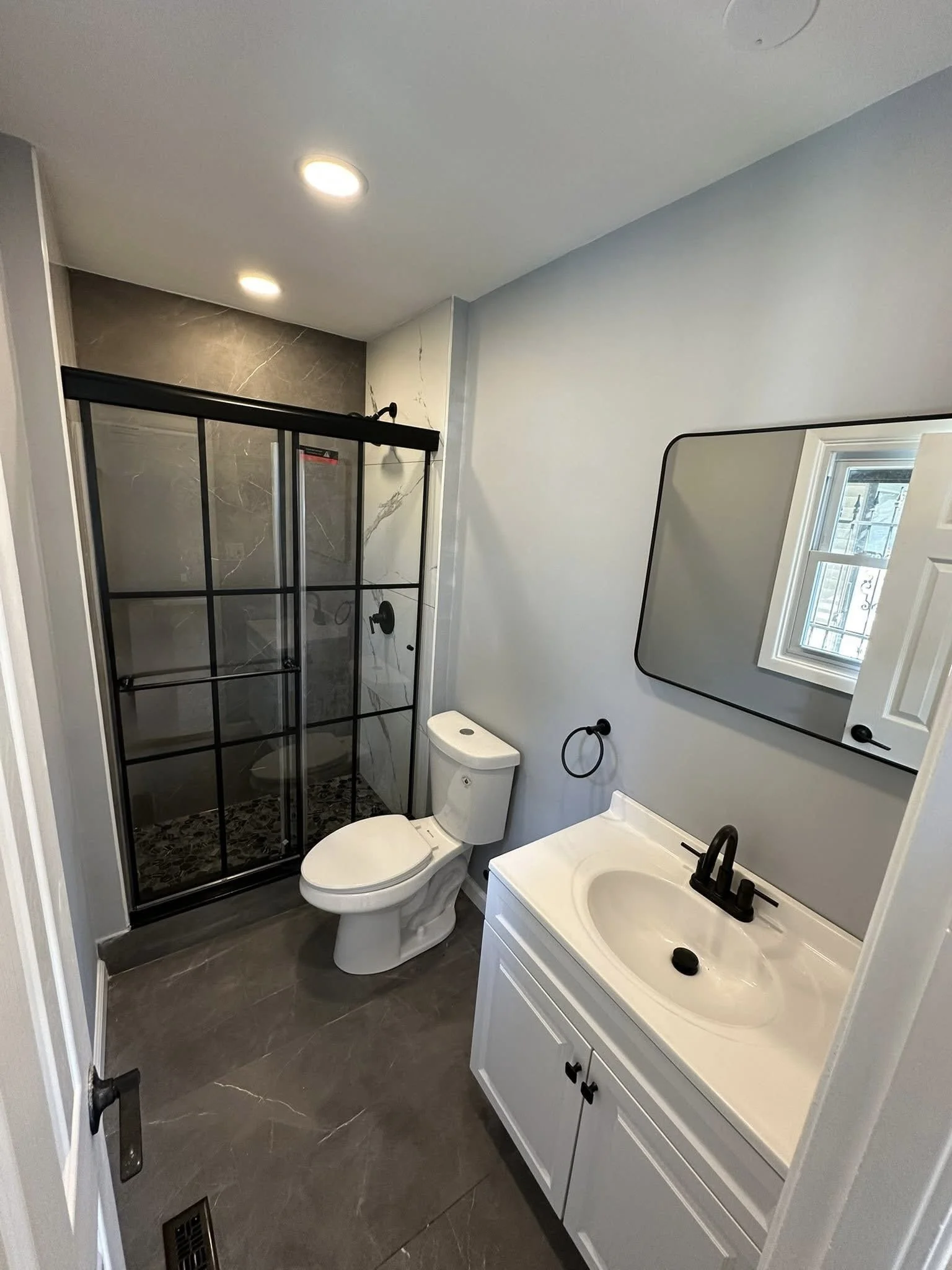 Modern bathroom with a walk-in shower with black framing and marble tiles, white toilet, white vanity with black fixtures, large mirror, and gray tile flooring.