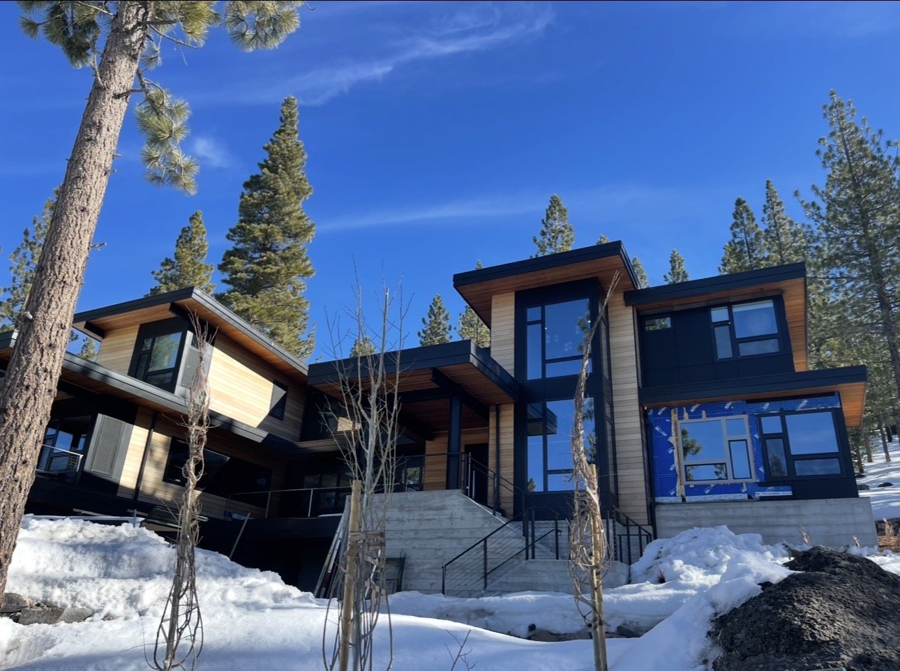 Modern house with large glass windows and wood siding, situated in a snowy landscape surrounded by pine trees under a clear blue sky.