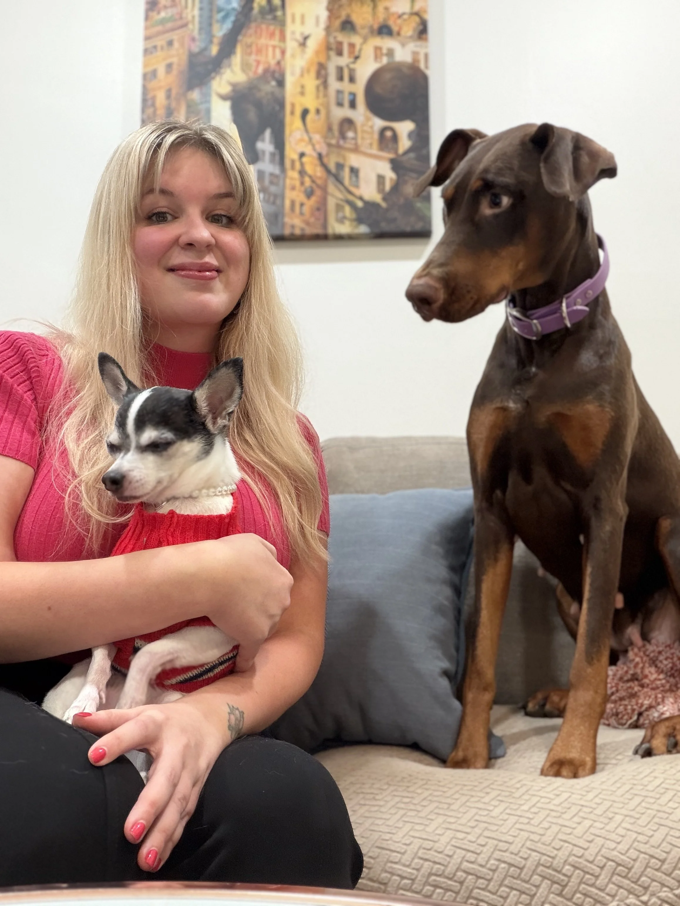 A woman with long blonde hair sitting on a beige couch, holding a small black and white dog wearing a red sweater, two dogs are visible: a small dog in her lap and a large Doberman sitting nearby on the couch, and a colorful abstract painting hanging on the wall behind them.