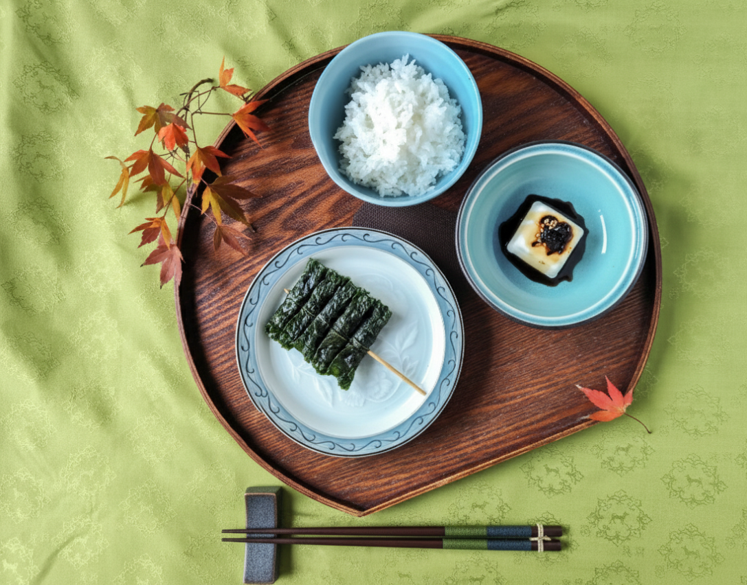 竹のトレイに乗った和風のお弁当セット。白米、海苔巻き、豆腐のおかきに醤油と黒ごまのソース。緑の楓の葉と箸が配置されている。