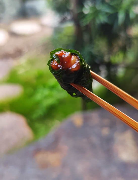 箸でつつまれた海苔巻き寿司、サーモンとイクラのトッピングが見える。背景は屋外の庭園の景色。
