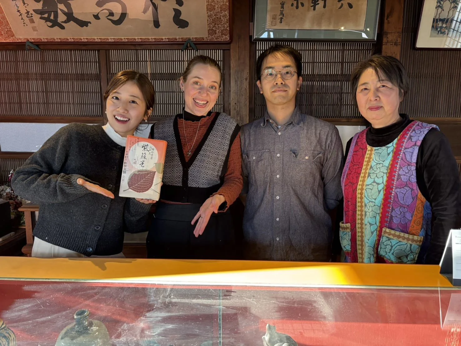 六軒京本舗の店内で、四人の男女が笑顔で写真を撮っている。左から女性、女性、男性、女性。左の女性は紙袋を持ち、右の女性は多色の刺繍入りのベストを着ている。背景には日本の書や絵が飾られている。