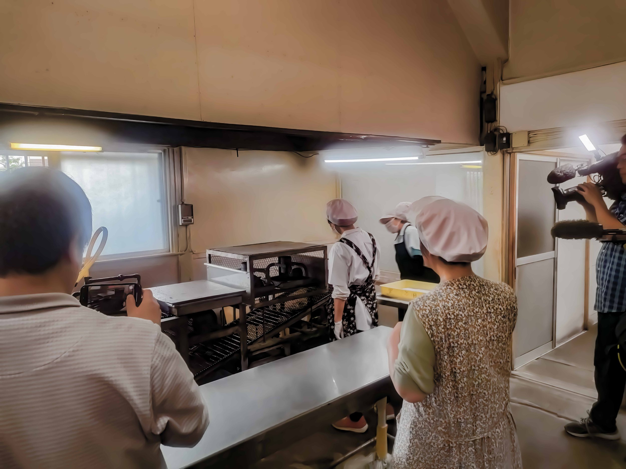 調理場で作業しているスタッフを取材するためのカメラマンと観客たち。調理場内にはステンレスの作業台や調理器具が見える。