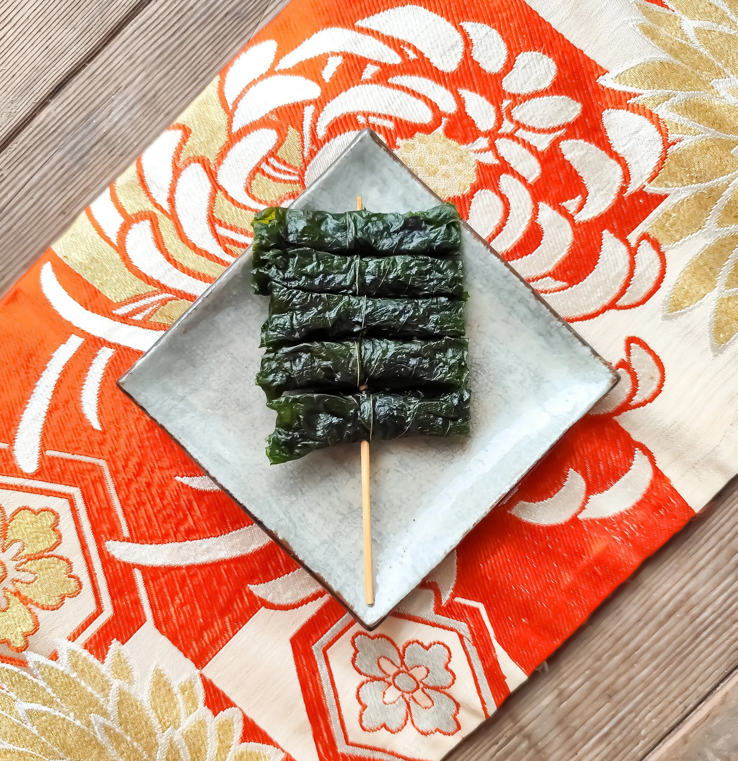 葉で包まれた伝統的な和菓子が竹串に刺さっている。背景は赤と金色の華やかな模様の布地。