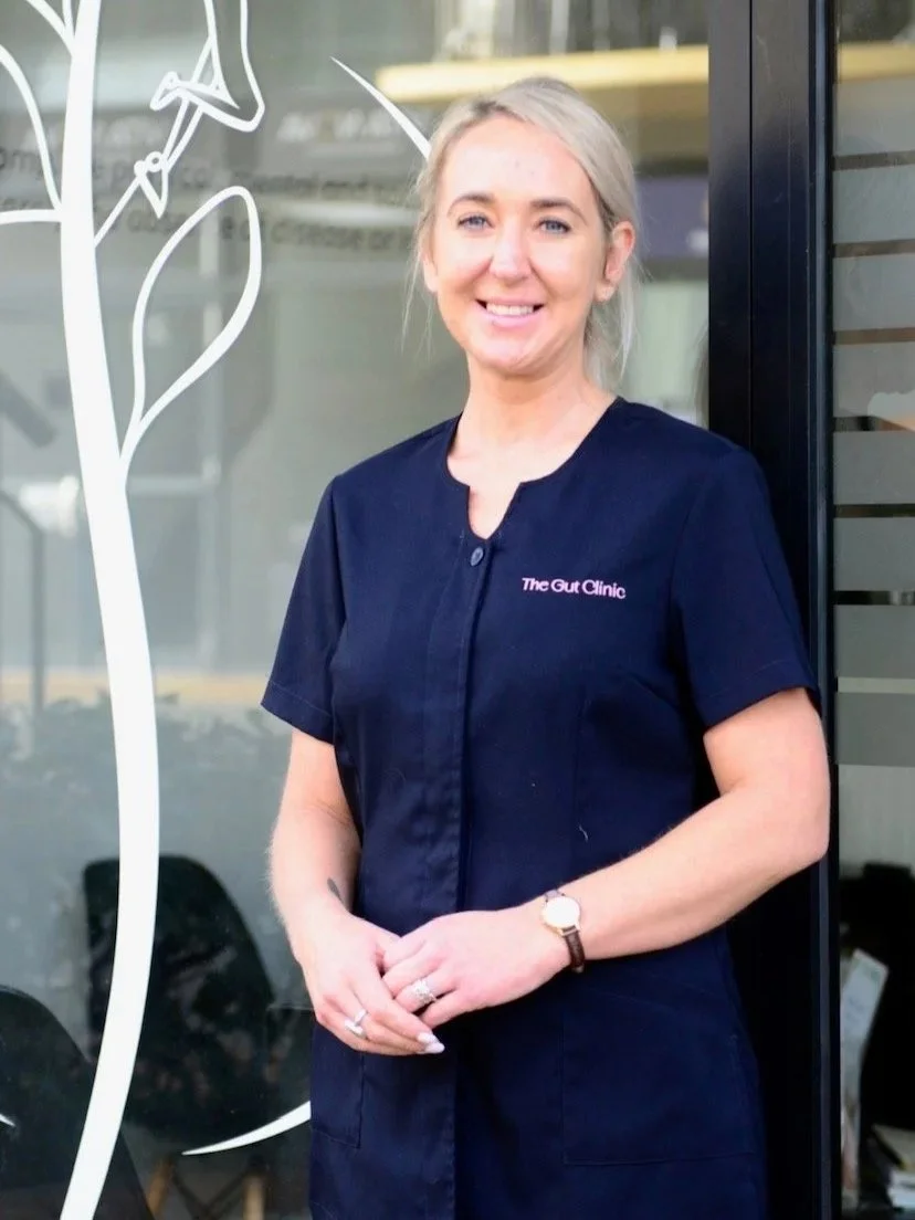 A woman with blonde hair in a blue uniform standing outside a building, smiling. The uniform has pink text that reads The Gut Clinic.