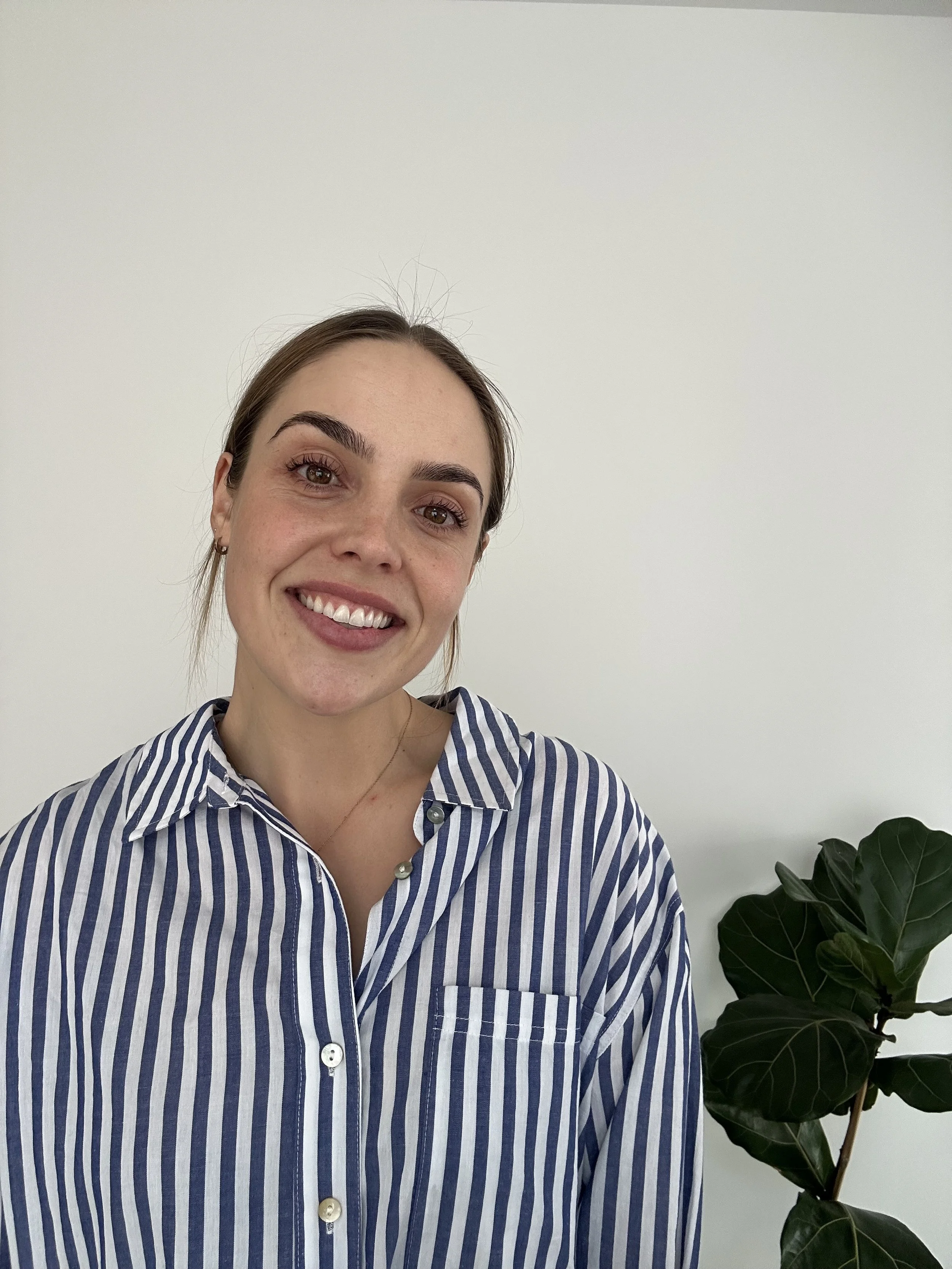 A smiling woman with brown hair and eyebrows, wearing a blue and white striped shirt, stands in front of a plain white wall and a potted dark green plant.