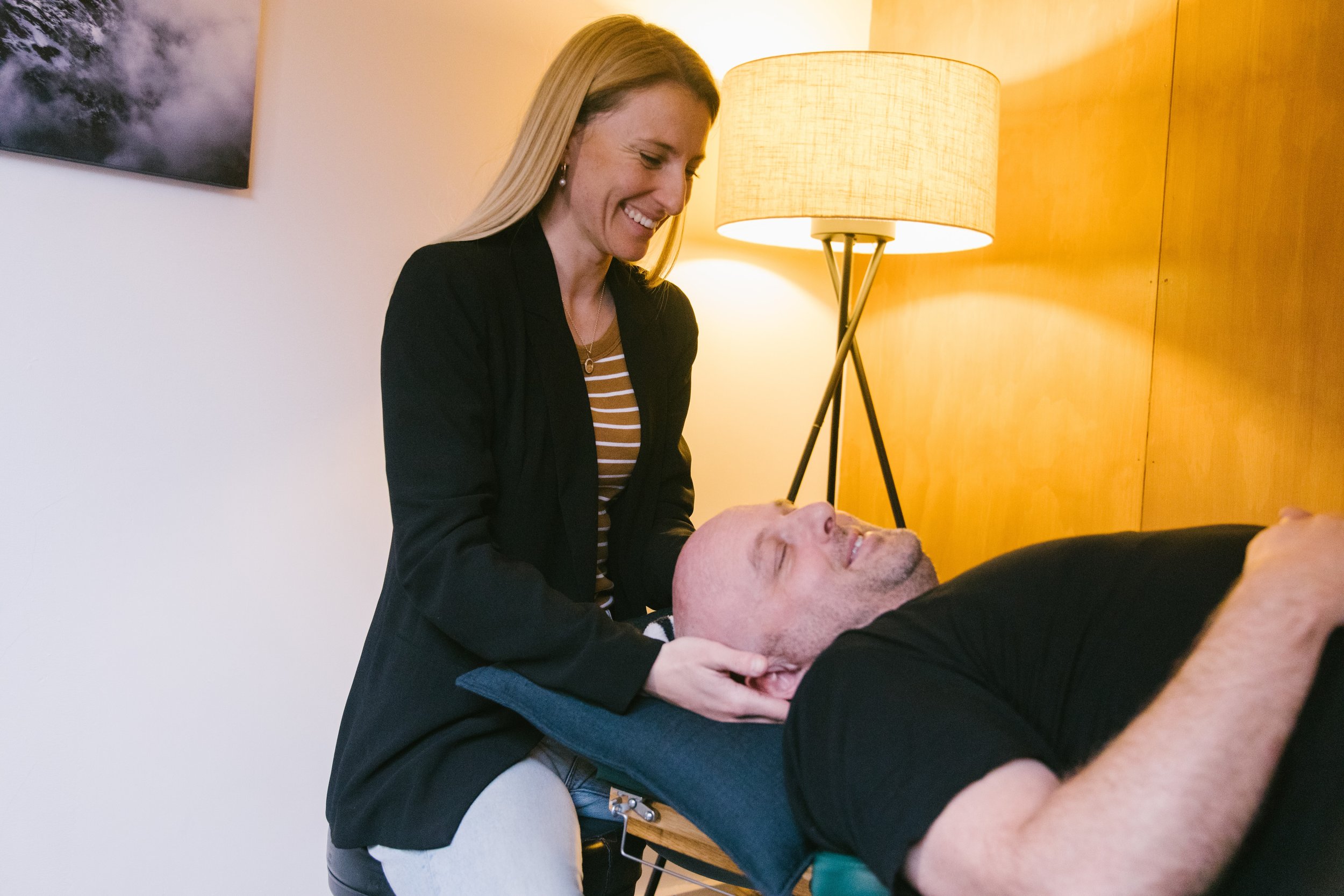 A woman giving a neck massage to a man who is lying on a massage table, smiling with his eyes closed, in a warmly lit room.