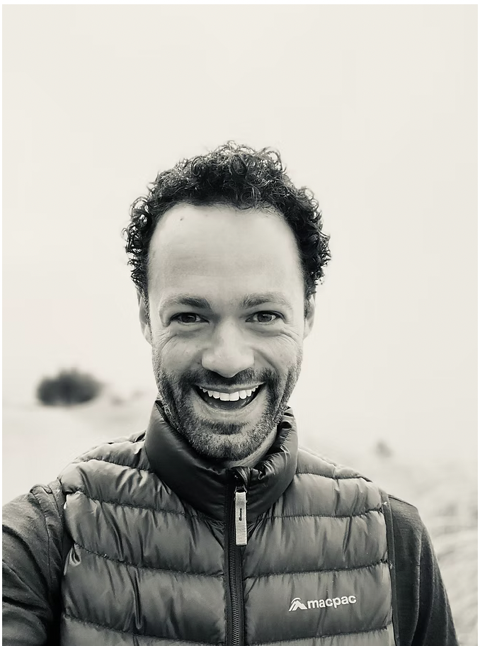 A man with curly hair and a beard smiling at the camera, wearing a puffer vest over a jacket outdoors.