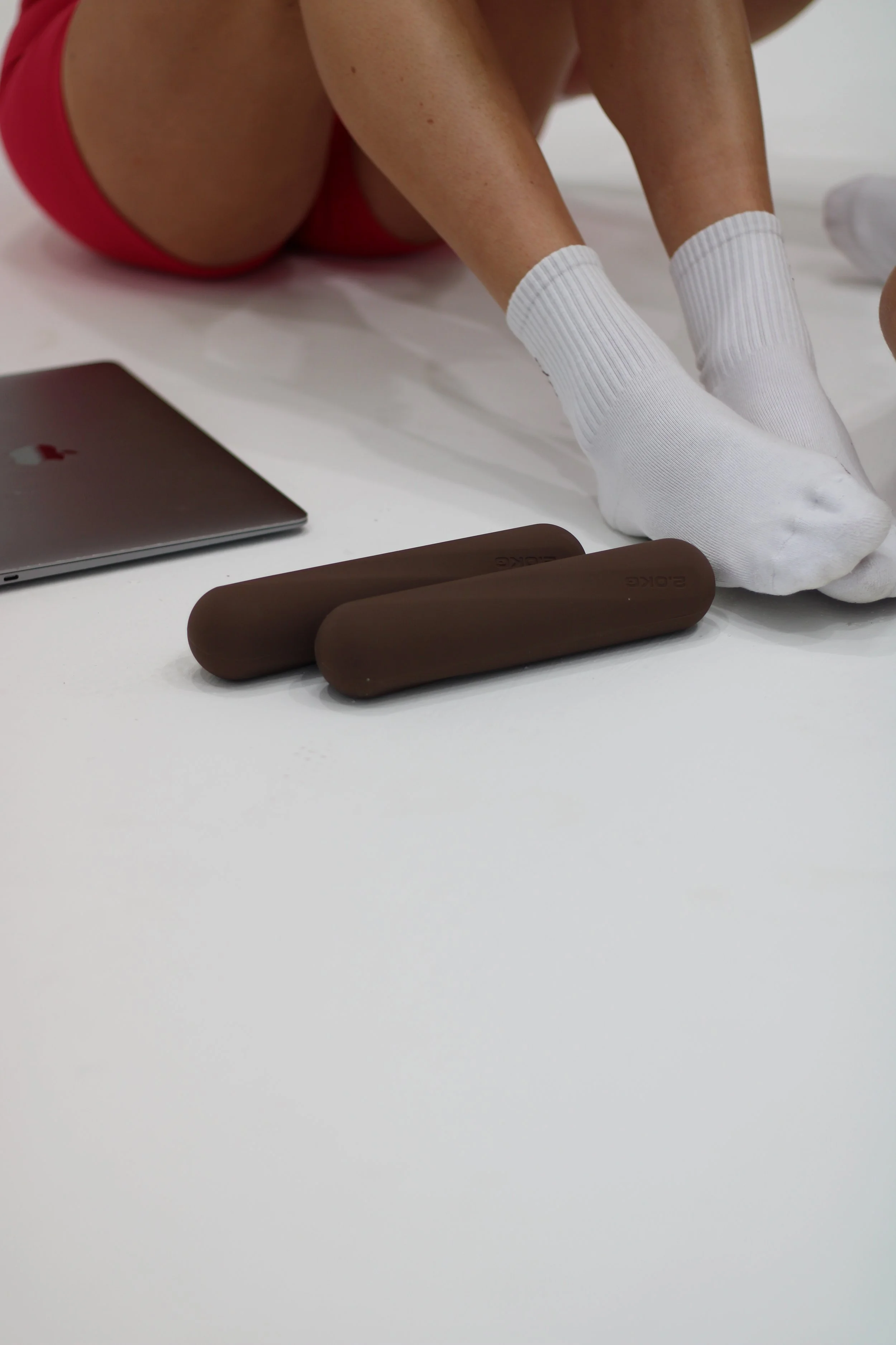Person sitting on the floor wearing white socks and red shorts with two dumbbells and a closed silver laptop nearby.