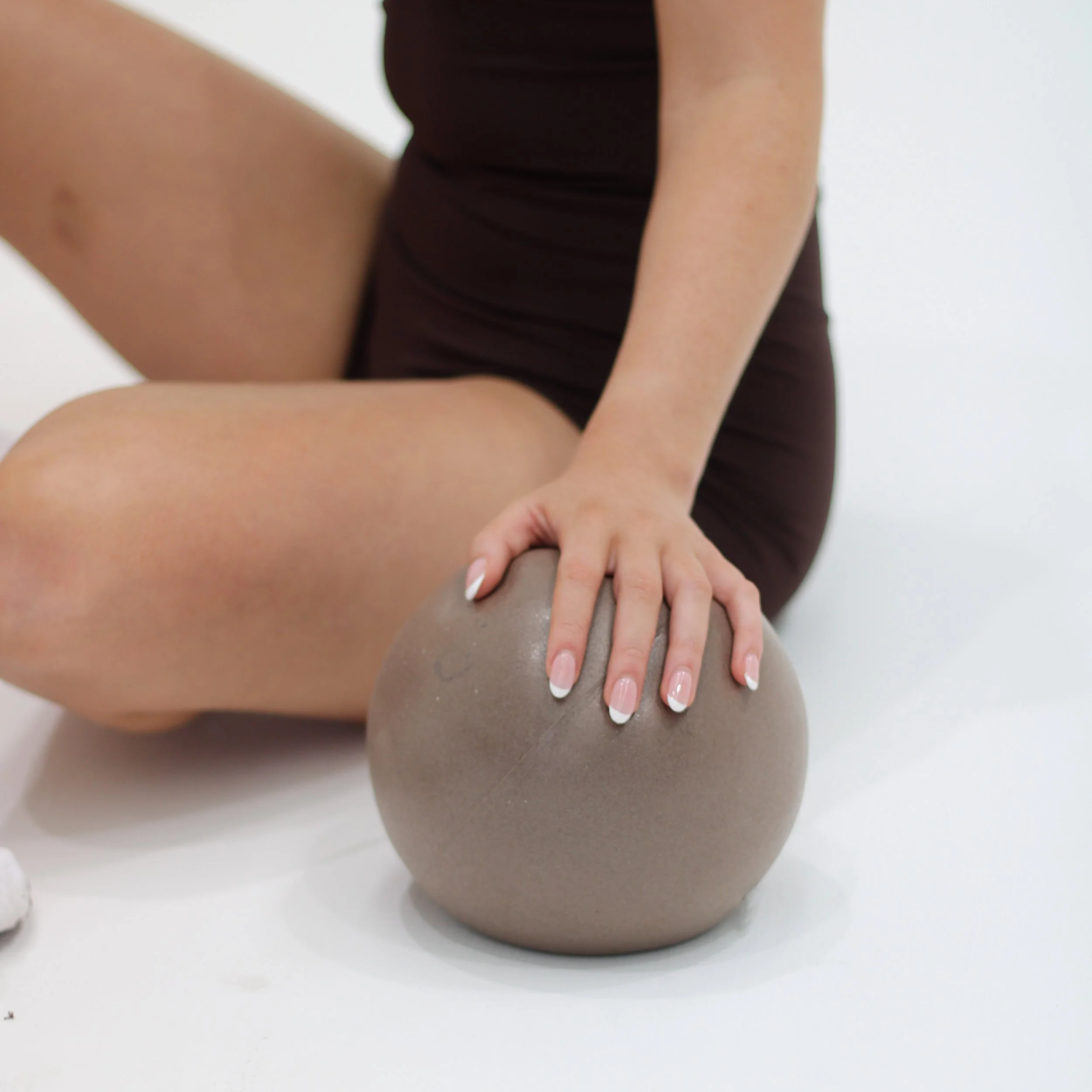 Person sitting on the floor with legs crossed with a Pilates ball, touching the Pilates ball with their right hand, wearing brown activewear.