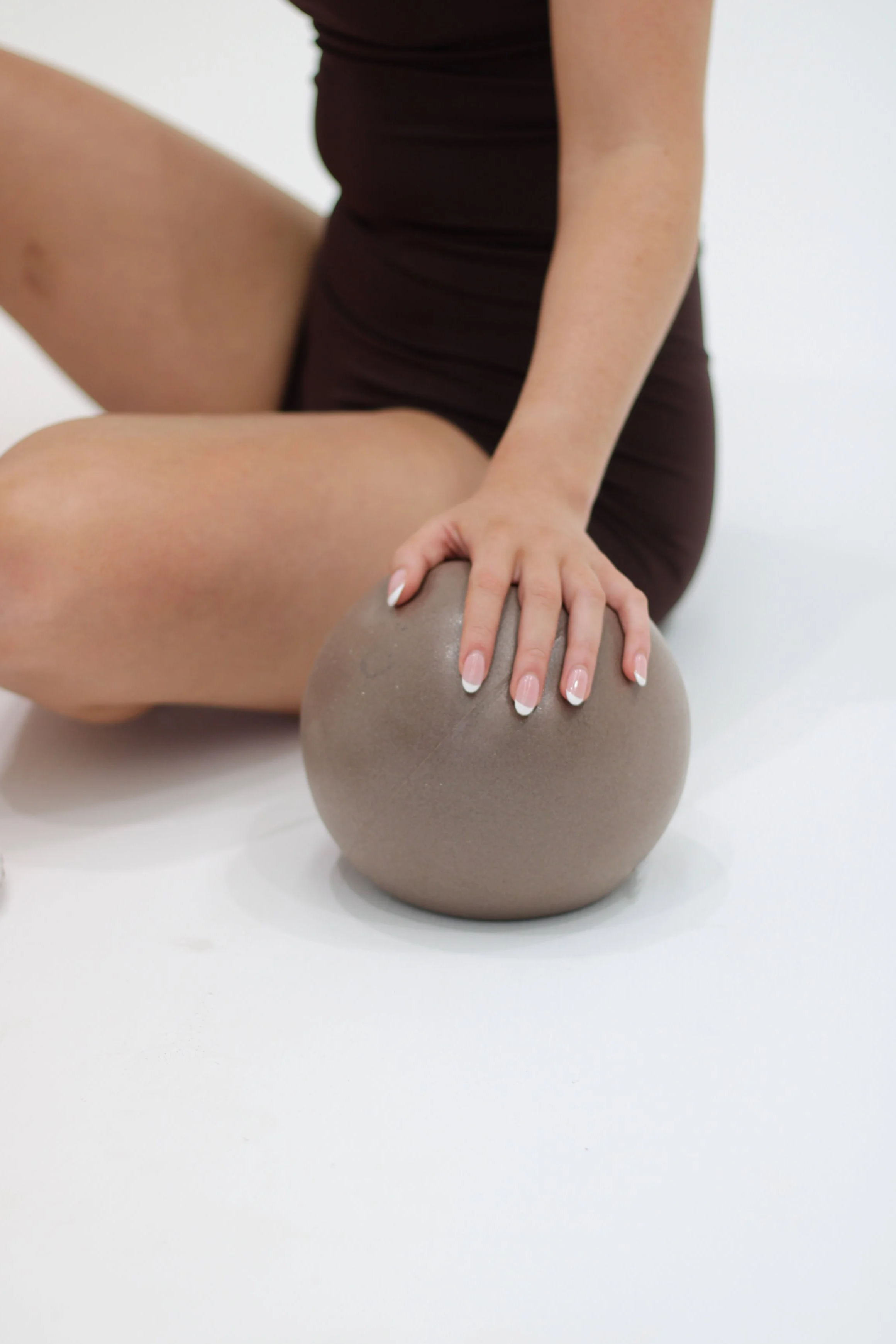 A person in a dark outfit is sitting on the floor, holding a Grey Pilates Ball