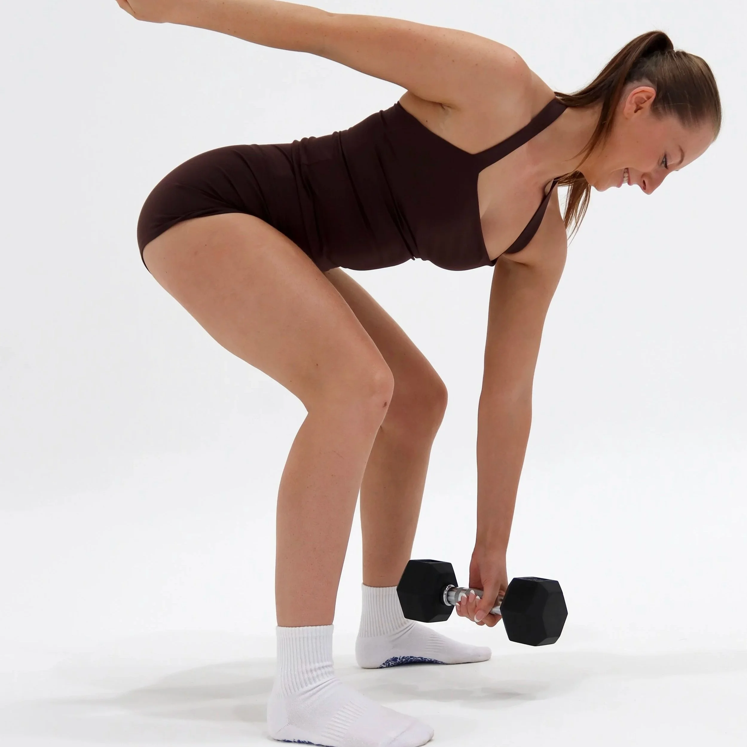 A woman in a black workout tank top, shorts, and white socks doing a bent-over dumbbell row exercise in a white studio.