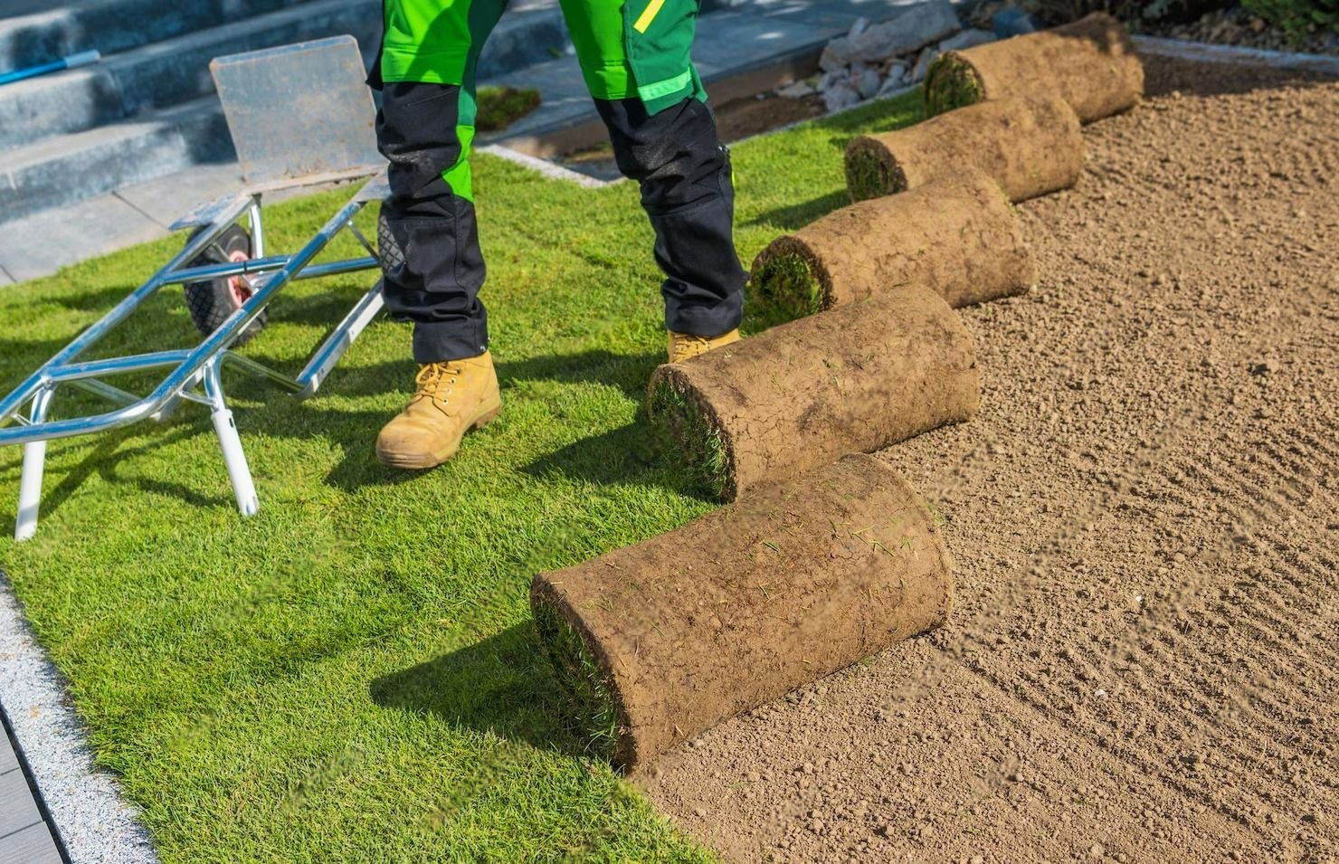 A worker in yellow boots and black and green pants uses a wheelbarrow on a grassy and sandy landscape with logs placed upright to create a border or edge.
