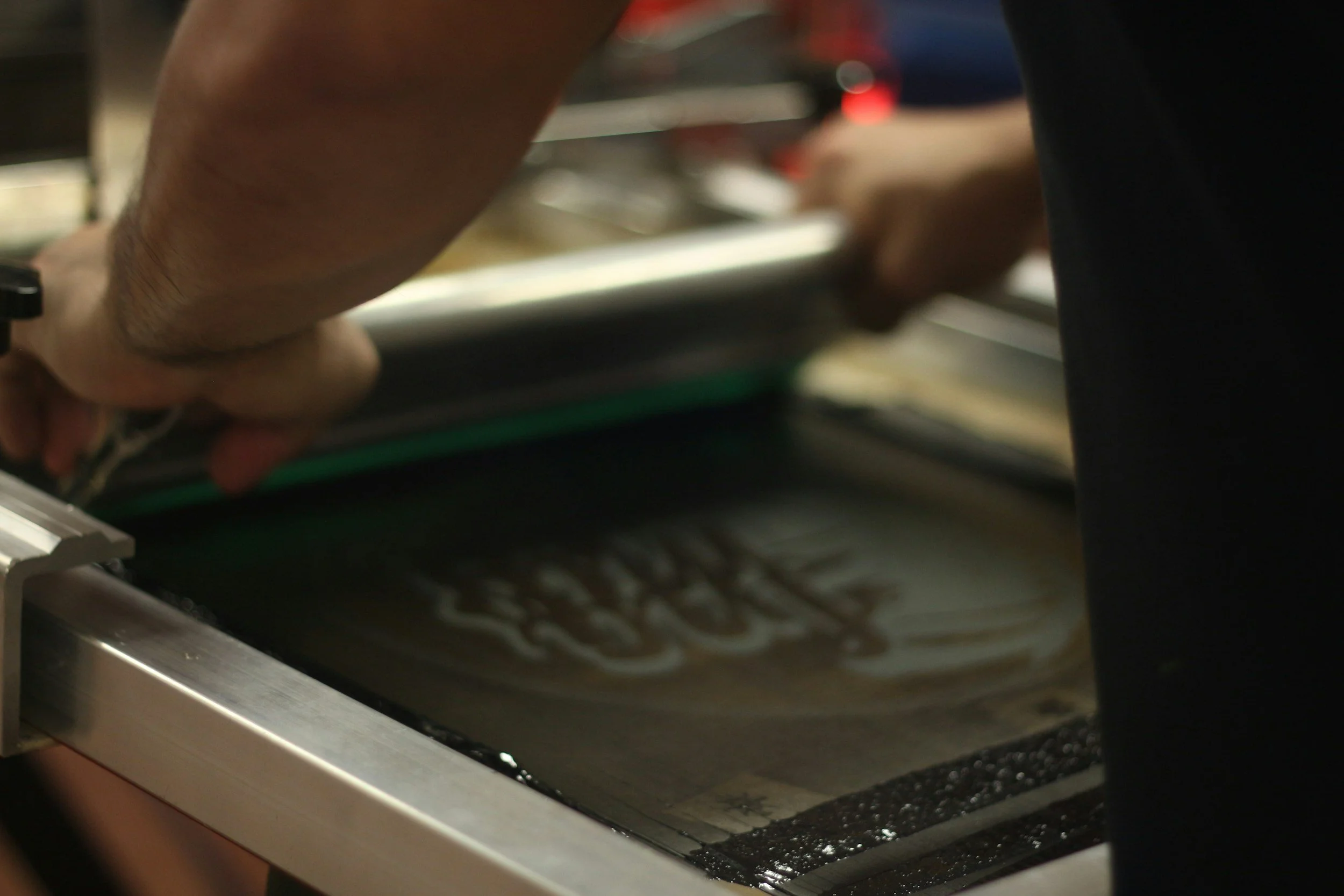 Close-up of person using a printing or heat press machine to print or transfer a design onto a dark-colored material, with visible metallic frame and machine components.