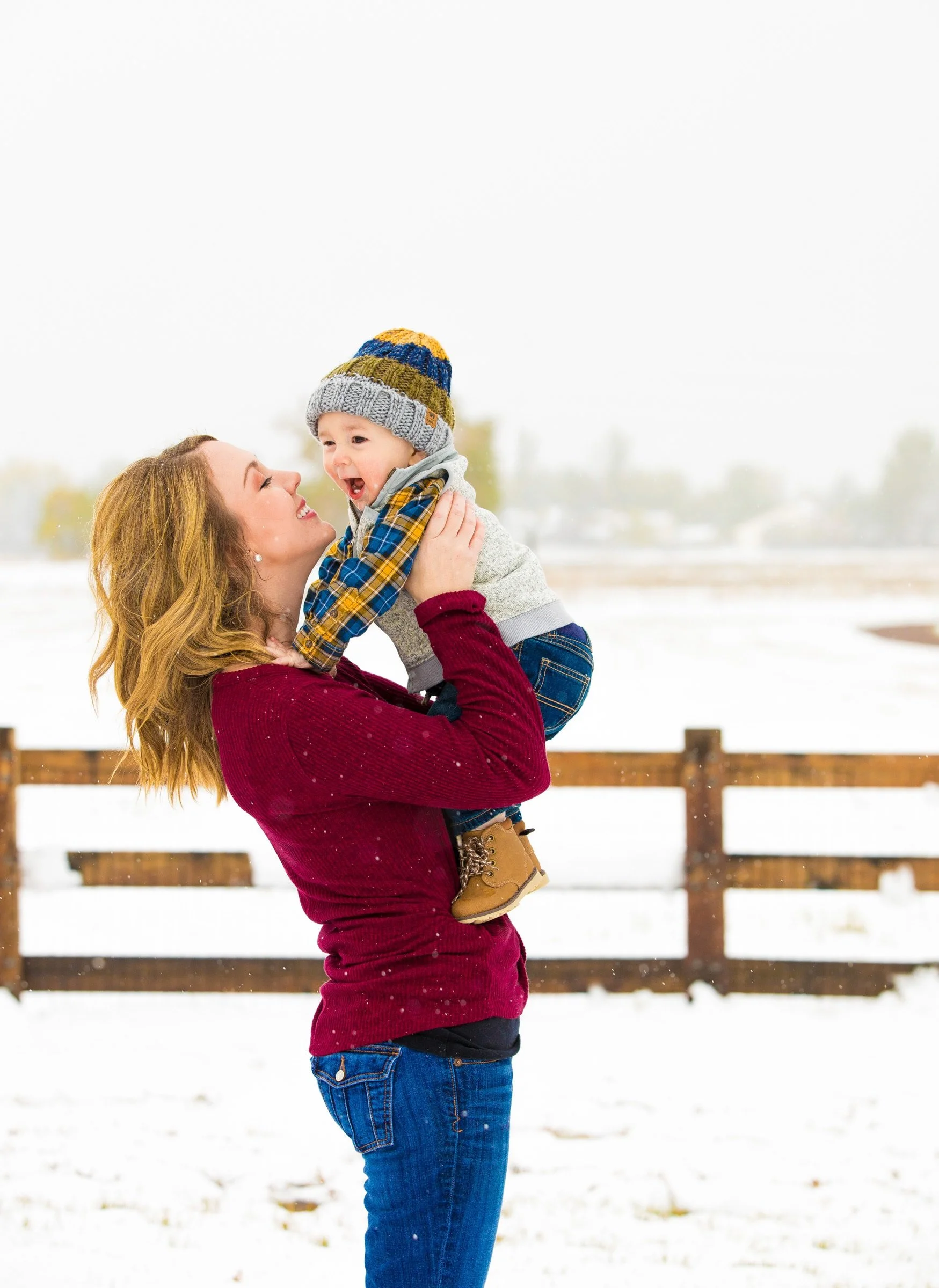 A woman holding a young boy up in the air outdoors in a snowy landscape, both smiling and looking happy.