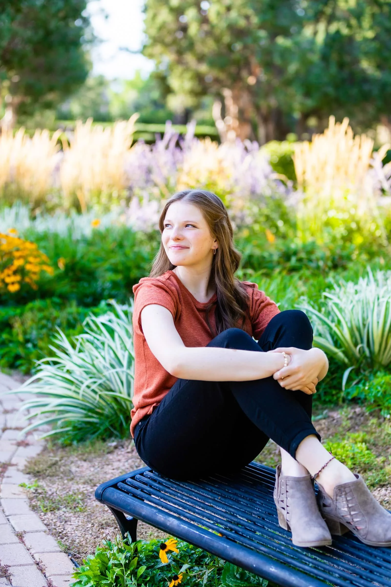 A young woman with long brown hair sitting on a black park bench in a garden filled with colorful flowers. She is wearing a rust-colored t-shirt, black pants, and beige ankle boots, with her arms wrapped around her knees, looking off to the side.