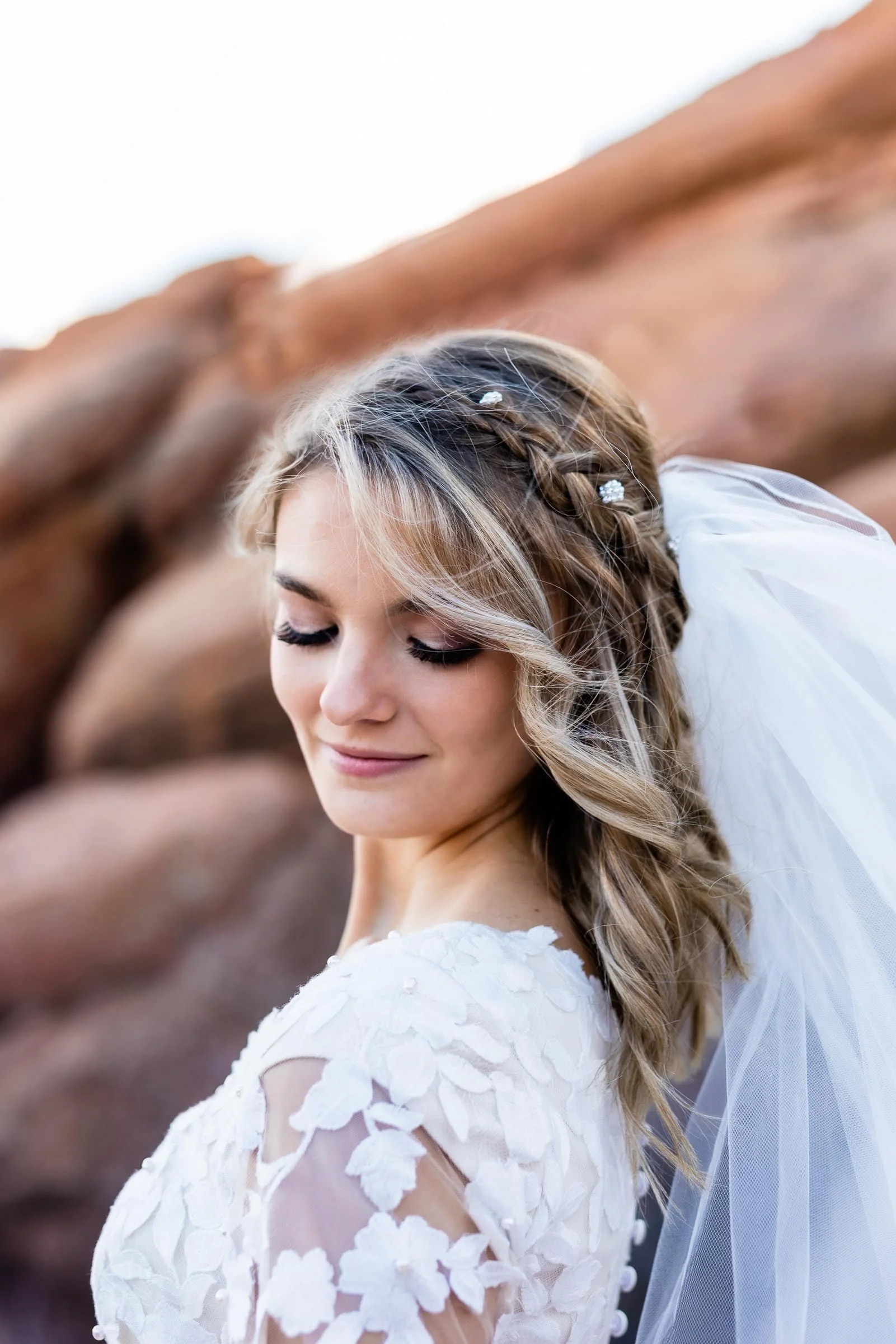 A bride with styled hair and a white lace wedding dress, standing outdoors near rocky terrain, with her eyes closed and a slight smile.