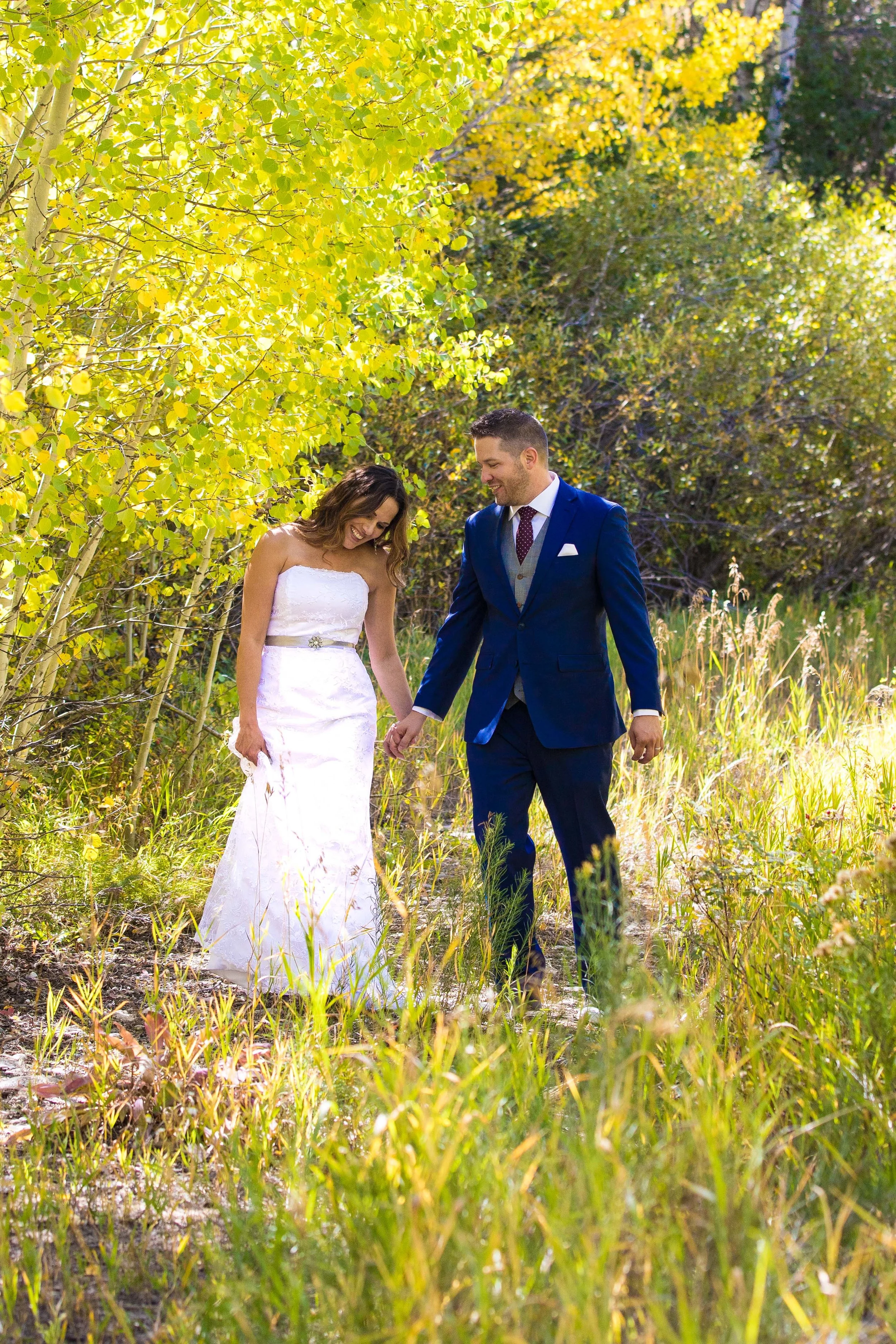 A bride and groom walking hand in hand through a sunlit forest, smiling and looking down at each other.