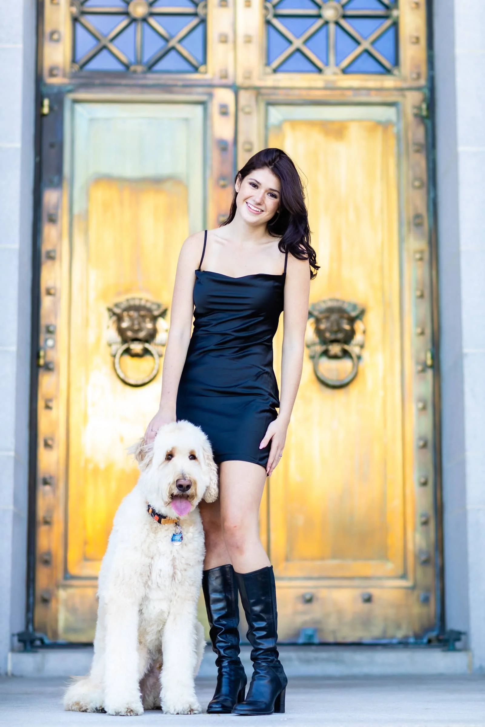 A woman in a black dress and knee-high black boots standing next to a large golden retriever with a pink tongue out, in front of a large ornate wooden door.