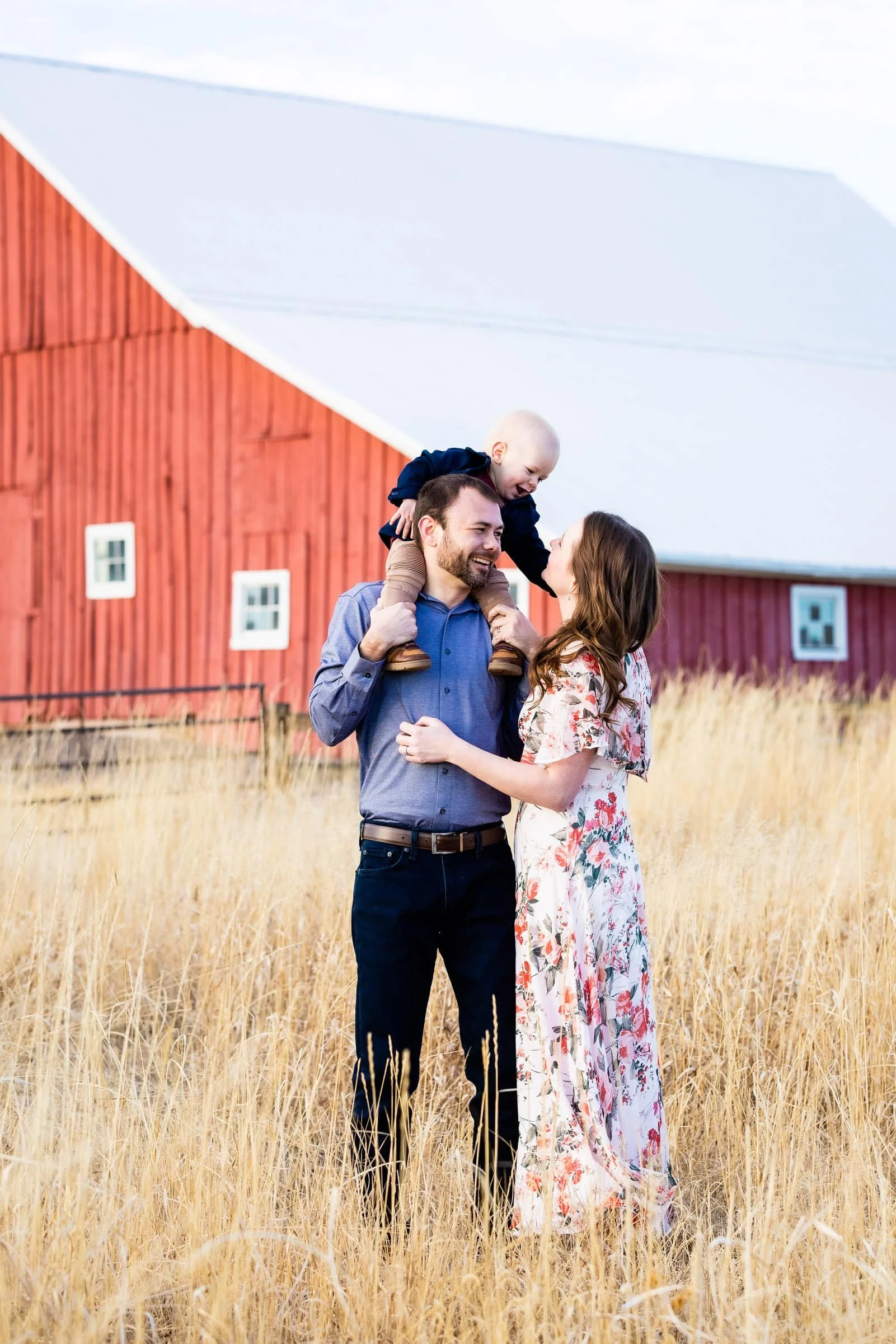 Family of three enjoying time together in a field with a red barn in the background, parents holding their young child on the father's shoulders.