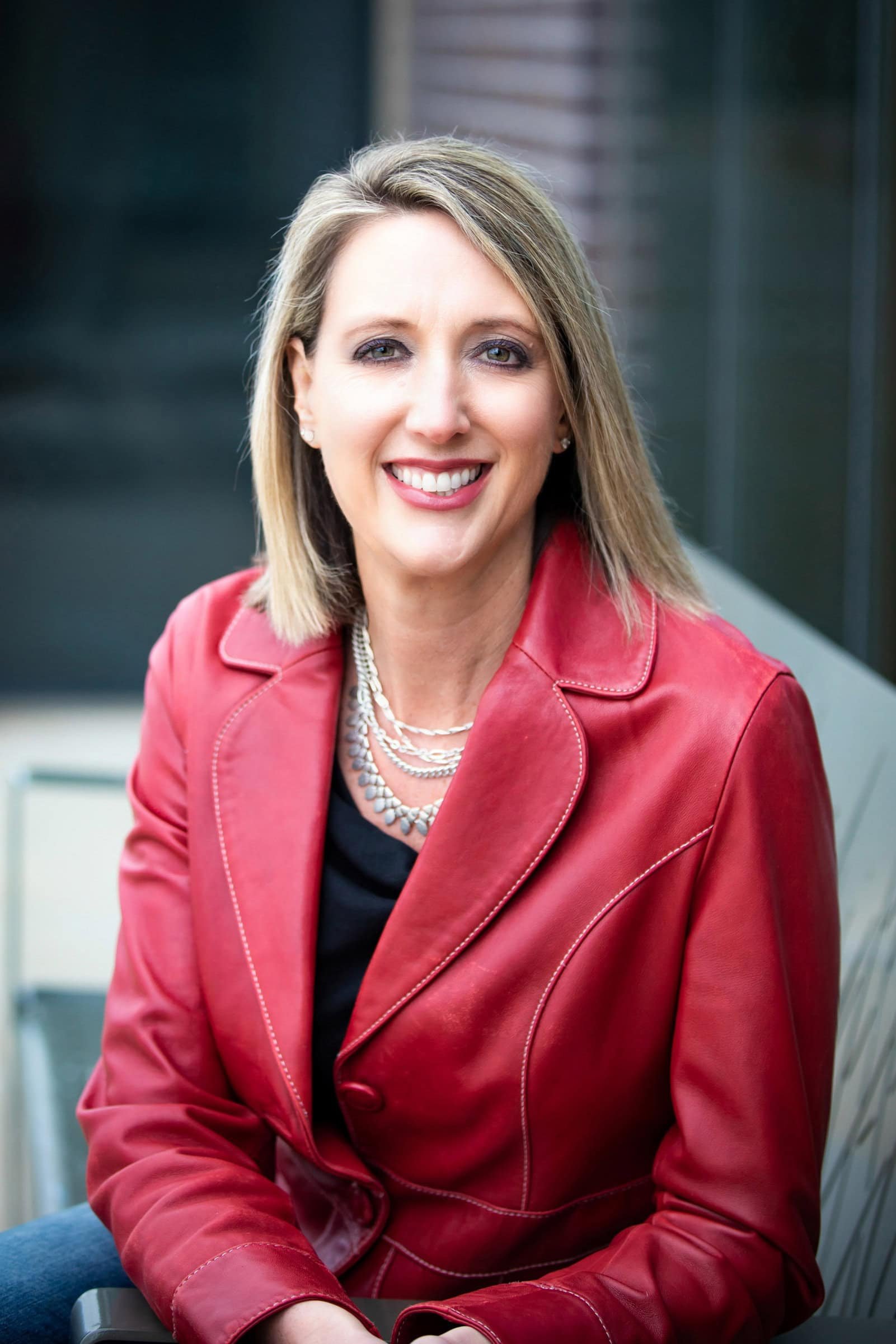 A woman with blonde hair, wearing a red leather jacket and a black top, sitting outdoors with a smile.