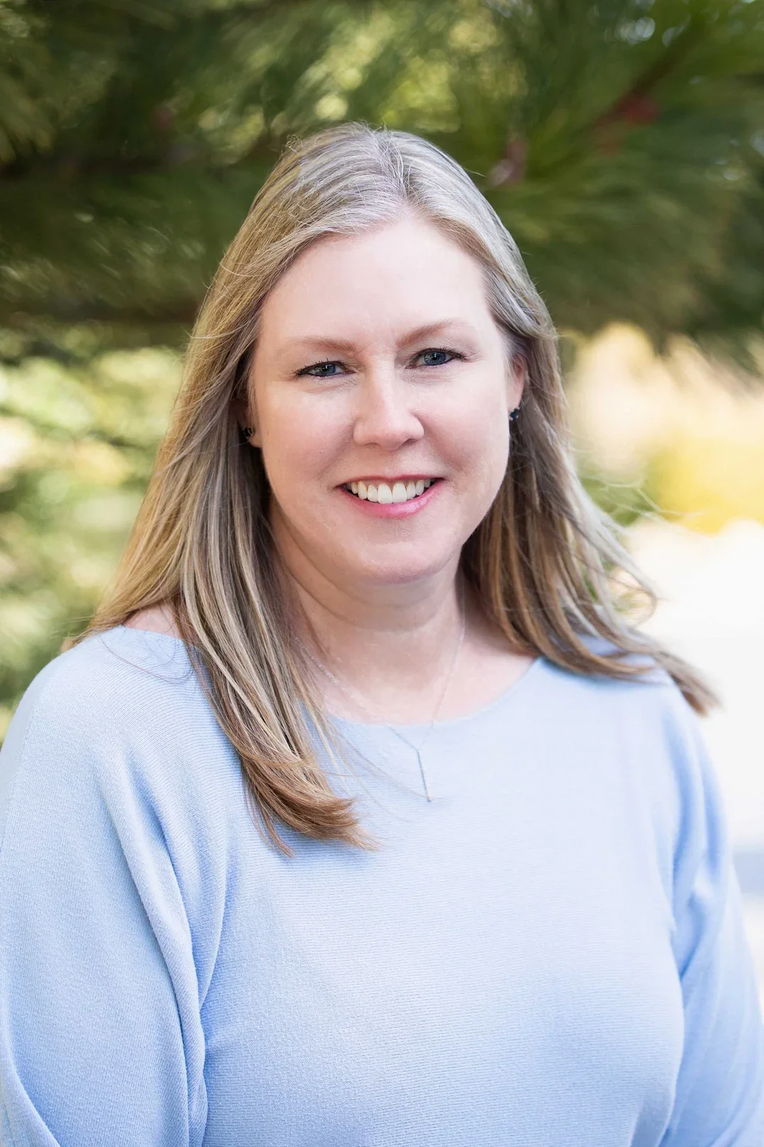 A smiling woman with shoulder-length blonde hair, wearing a light blue top, outdoors with green foliage background.